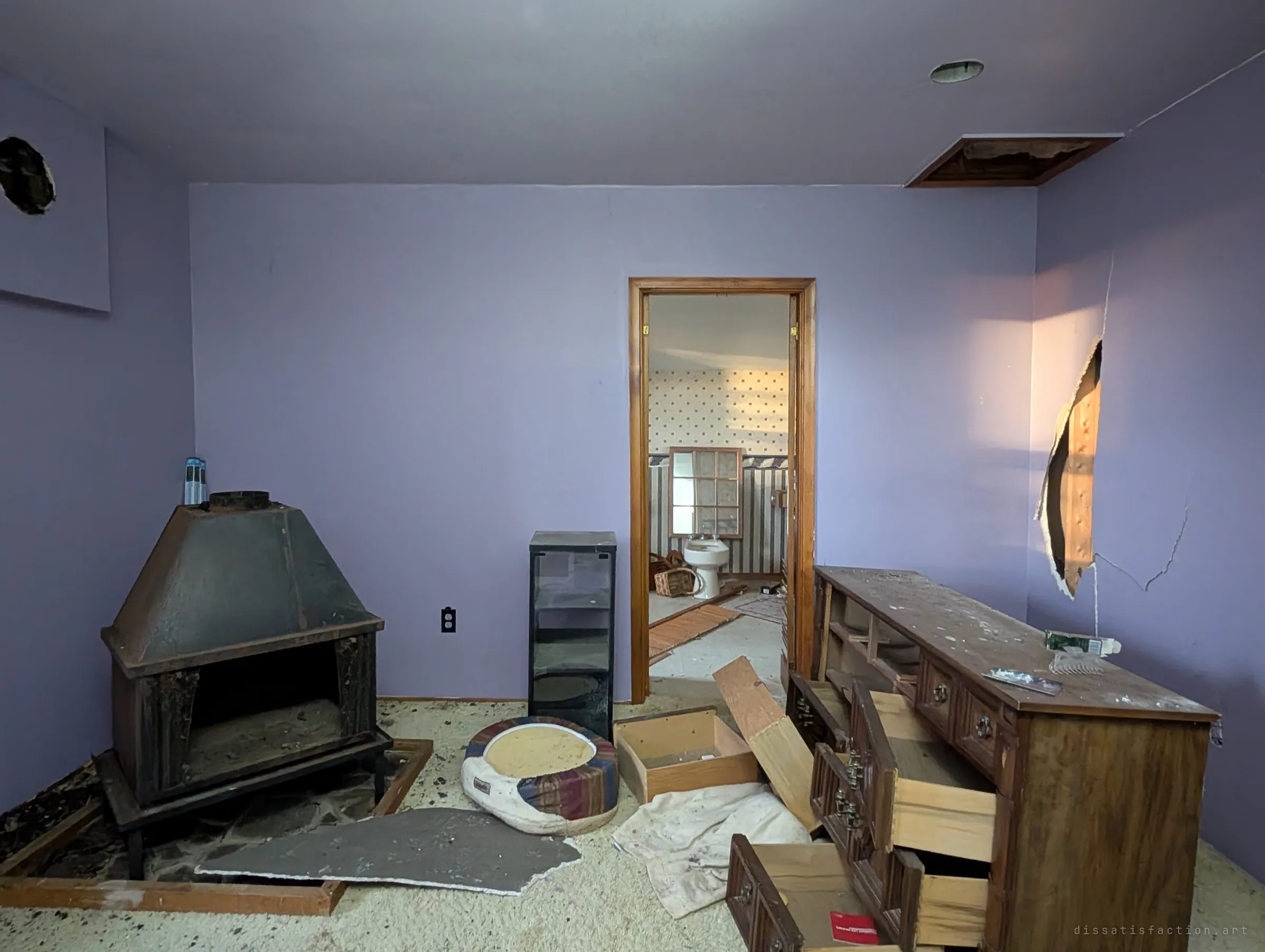 A lavender bedroom with a black freestanding wood stove, a pulled-apart dresser and holes punched in the walls, a wallpapered bathroom visible through the doorway. Soft afternoon light on the walls contrasts with the damage.