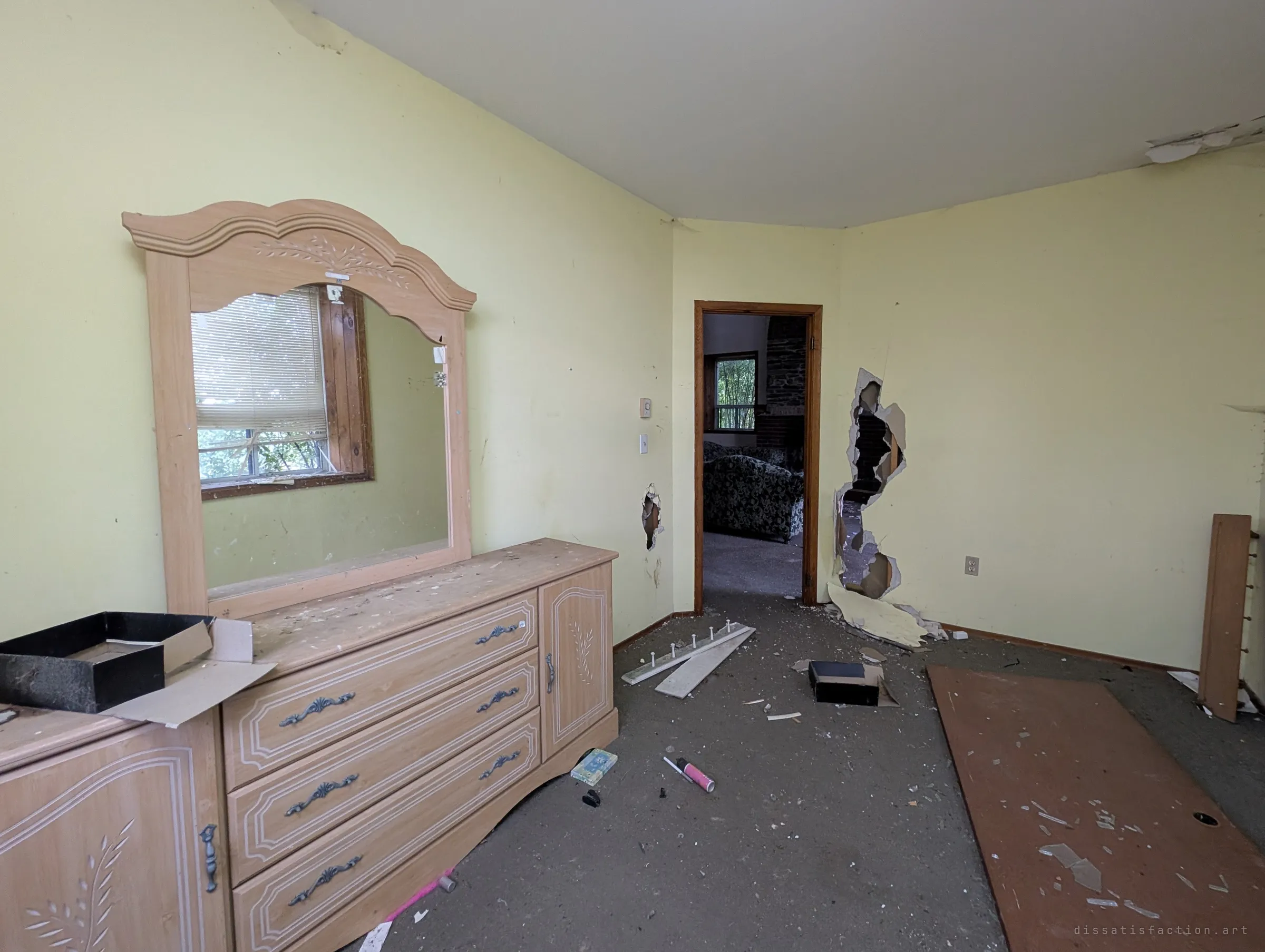 A pale yellow bedroom with a dresser and arched mirror, drywall torn open beside the doorway that looks back onto the living room sofas. The broken walls show how vandals have moved through the house.