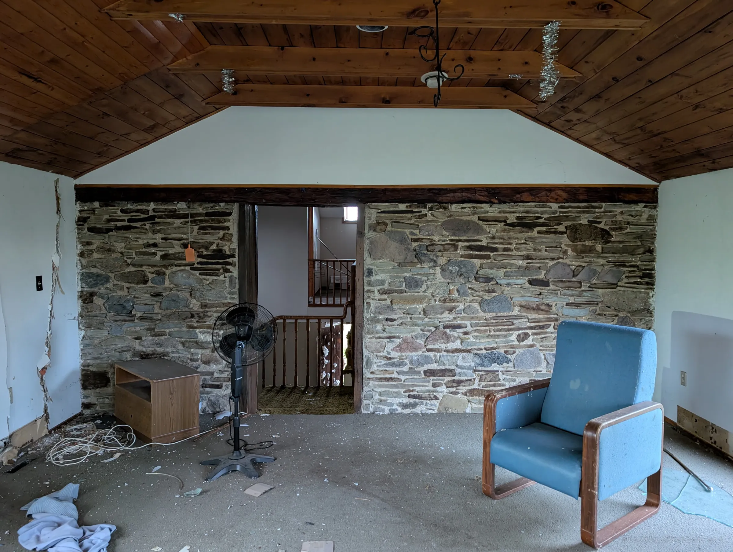 An attic room under a pine ceiling with an exposed fieldstone wall, a blue armchair and a standing fan, a doorway opening onto the stair landing. The old stone core of the house shows through the later renovation.