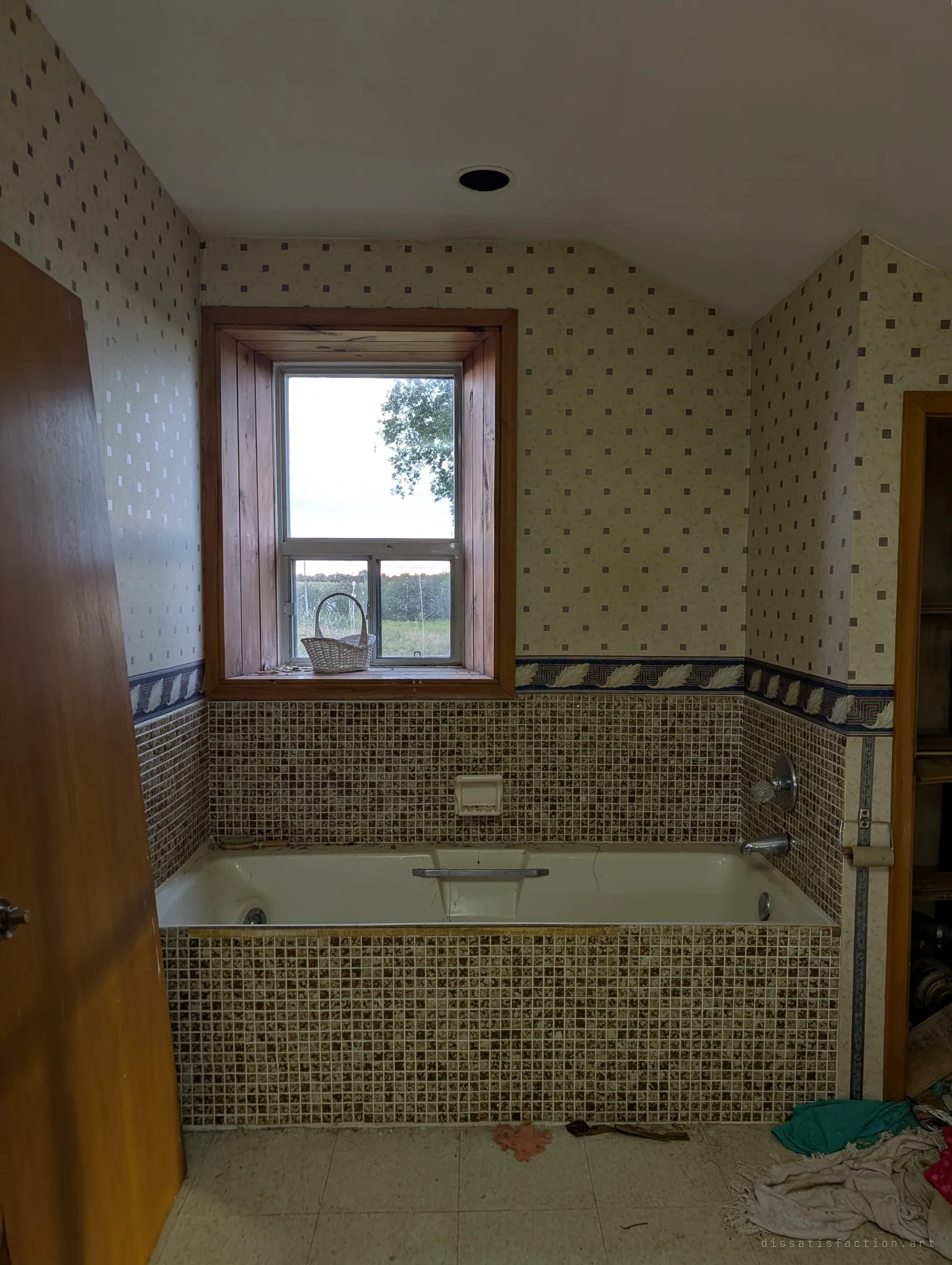 An upstairs bathroom under a sloped ceiling, a tiled tub set below a single window with a wicker basket left on the sill. Dated patterned wallpaper and scattered towels show a home abandoned mid-life.