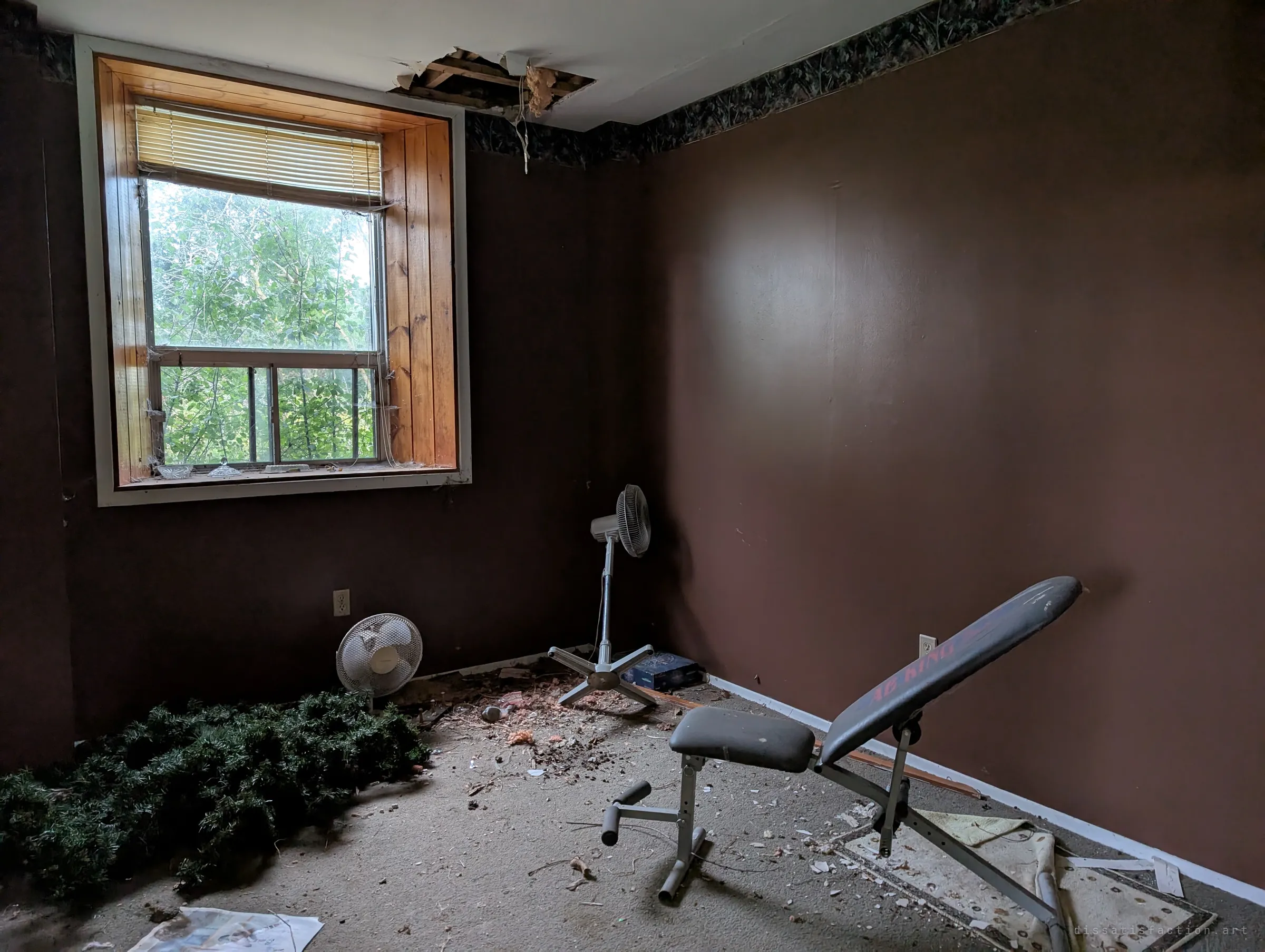 A dark brown bedroom with a hole torn in the ceiling, a weight bench, two fans and a heap of artificial garland on the debris-strewn carpet. Green leaves press against the window, showing how far the grounds have grown in.