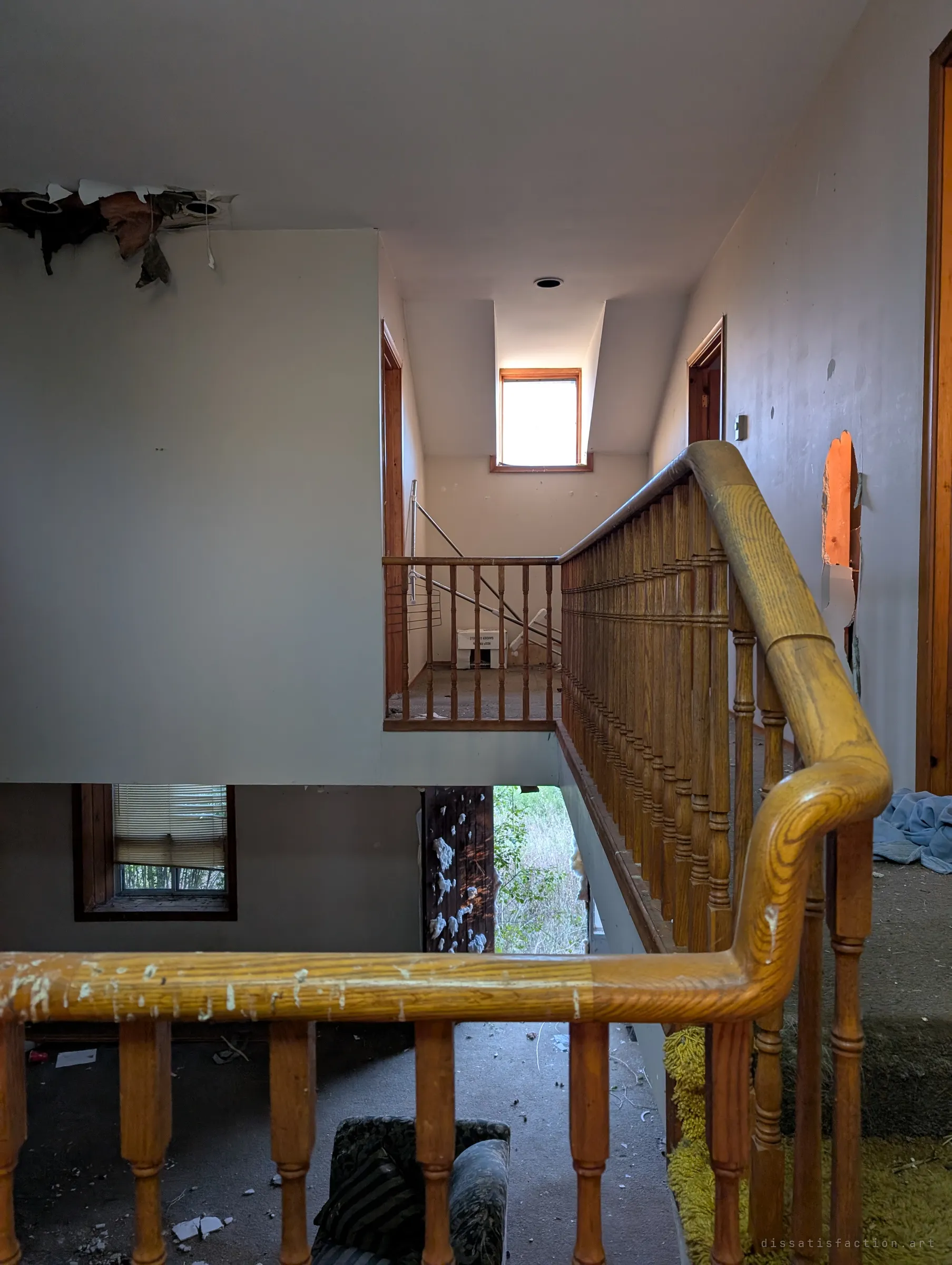 Looking across a wooden banister from the upstairs landing toward a gallery and a small bright window, with torn ceiling and punched walls. Below, an open doorway lets the overgrown yard spill light into the stairwell.