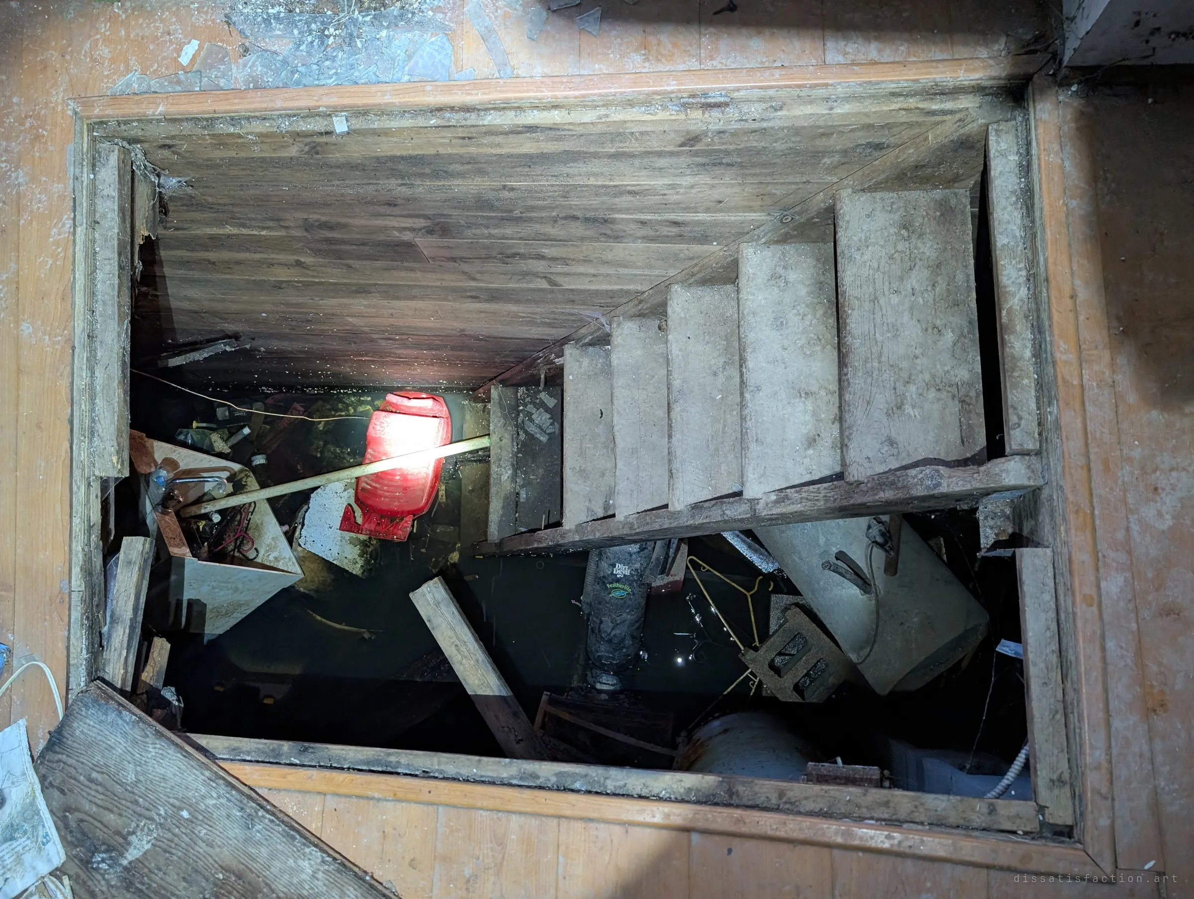 Looking down through an open floor hatch at a steep wooden stair descending into a flooded cellar, a red lamp glowing above black water and floating debris. The drowned basement is one of the most unsettling corners of the house.