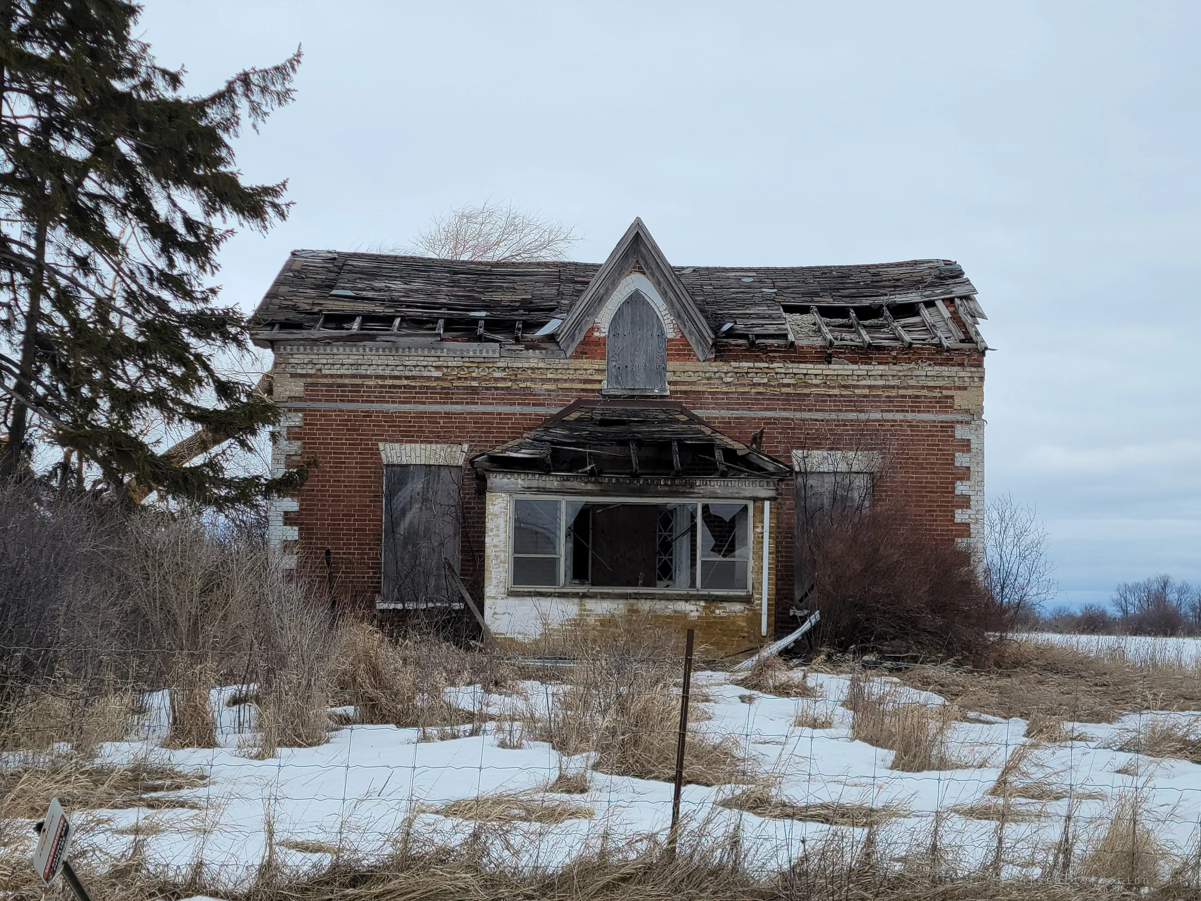 The front of an abandoned brick farmhouse with a pointed centre gable, its roof caving in and bay window smashed, behind a wire fence in snow. It sets the scene for the whole farmstead.