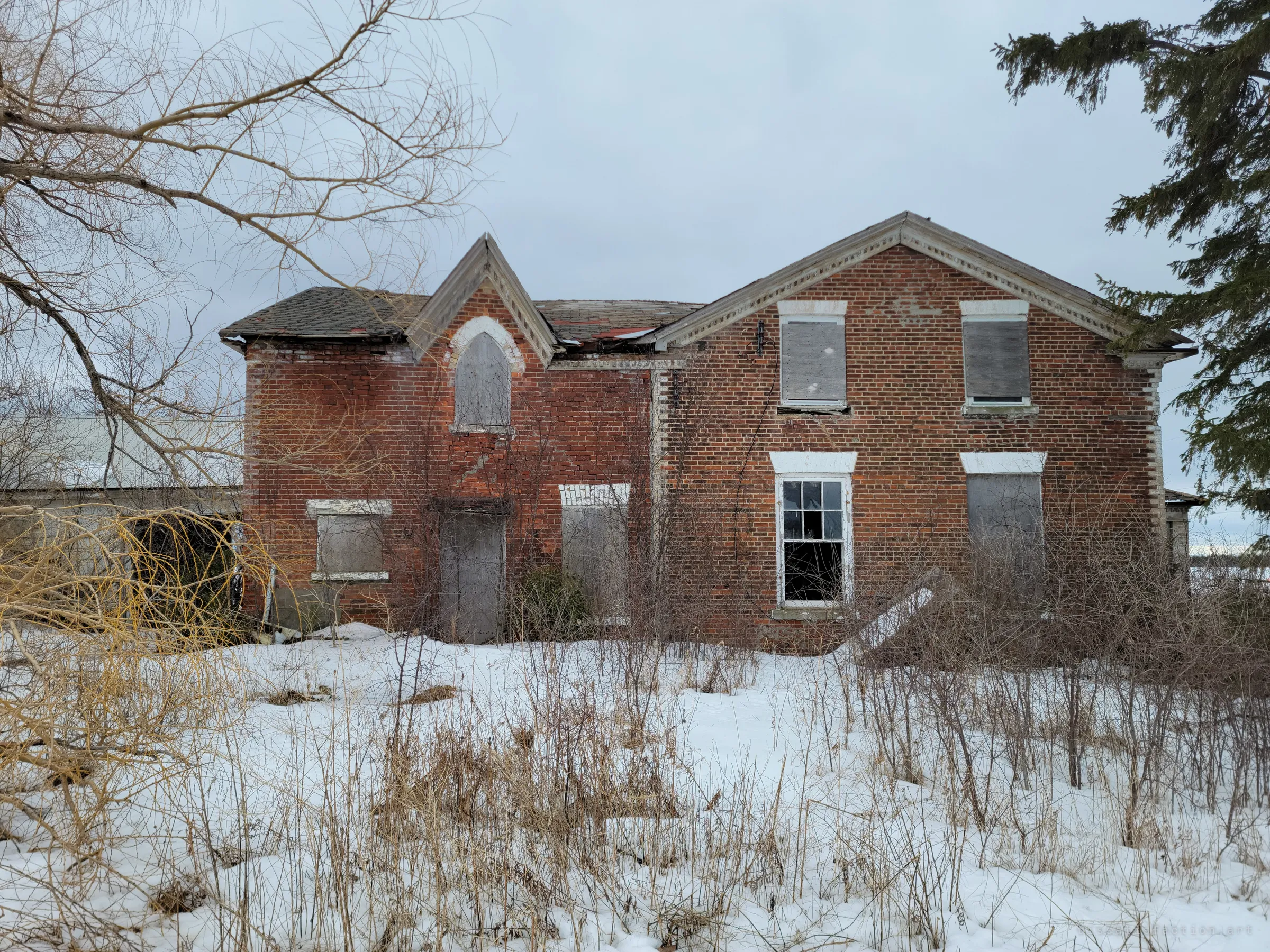 Another side of the brick farmhouse, most windows boarded over and bare shrubs grown up the walls, with deep snow in front. The one open window with broken panes hints at the interior.