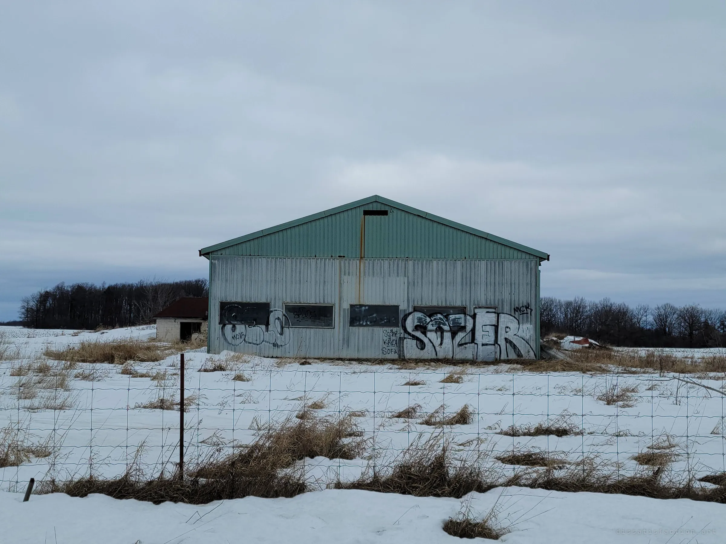 A green-roofed steel barn covered in graffiti stands alone in a snowy field behind a wire fence. The tags show others have found the property before.