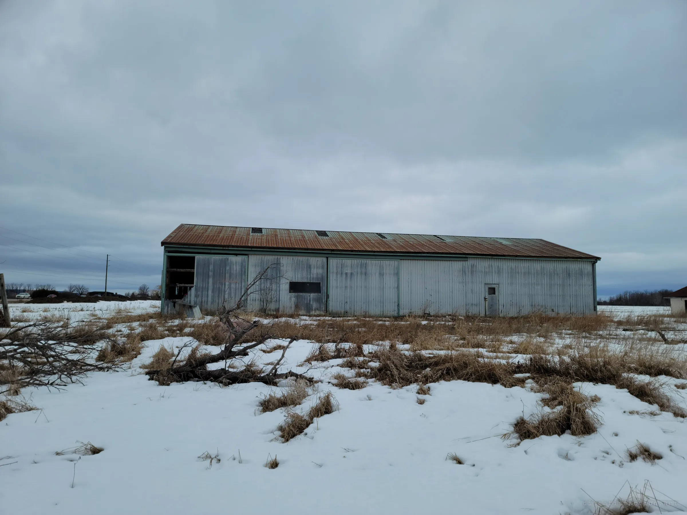 A long steel barn with a rusted roof stretches across a flat snowy field under heavy cloud. A fallen tree lies in the foreground grass.
