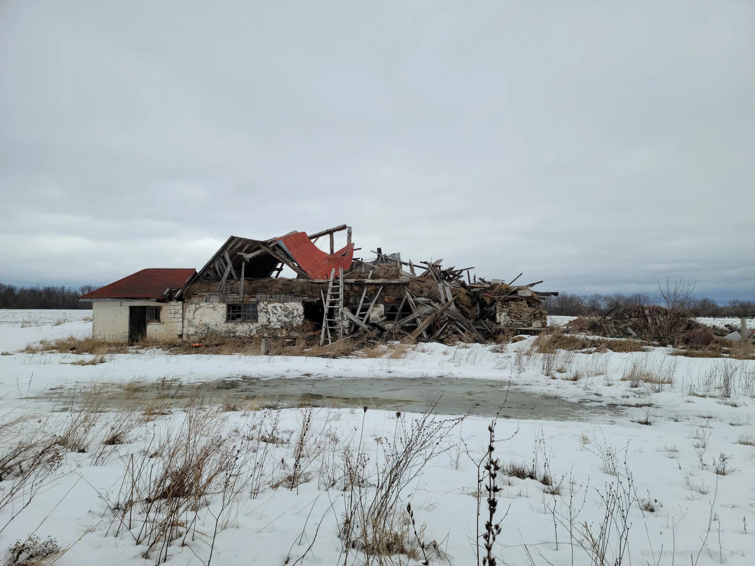 A collapsed barn beside a small block outbuilding, its red metal roof peeled up and timbers heaped in a pile, with a frozen pond in front. The farm is visibly falling apart.
