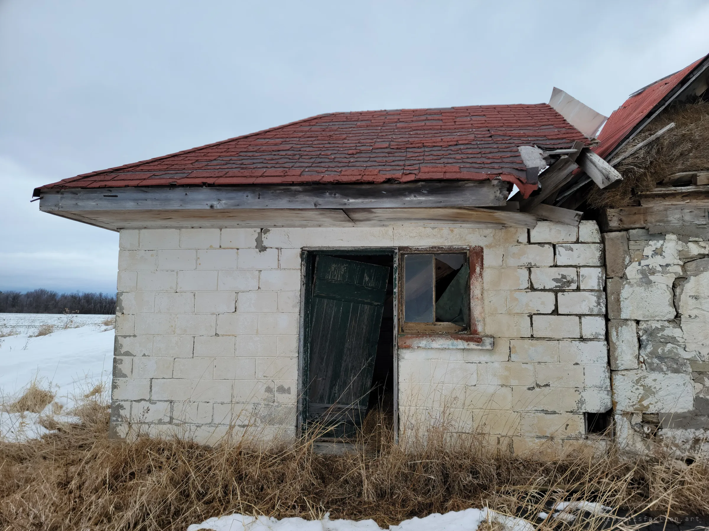 A whitewashed concrete-block outbuilding with a red shingle roof, a green door hanging off its hinges and a broken window. It joins the collapsing stone barn at the right.