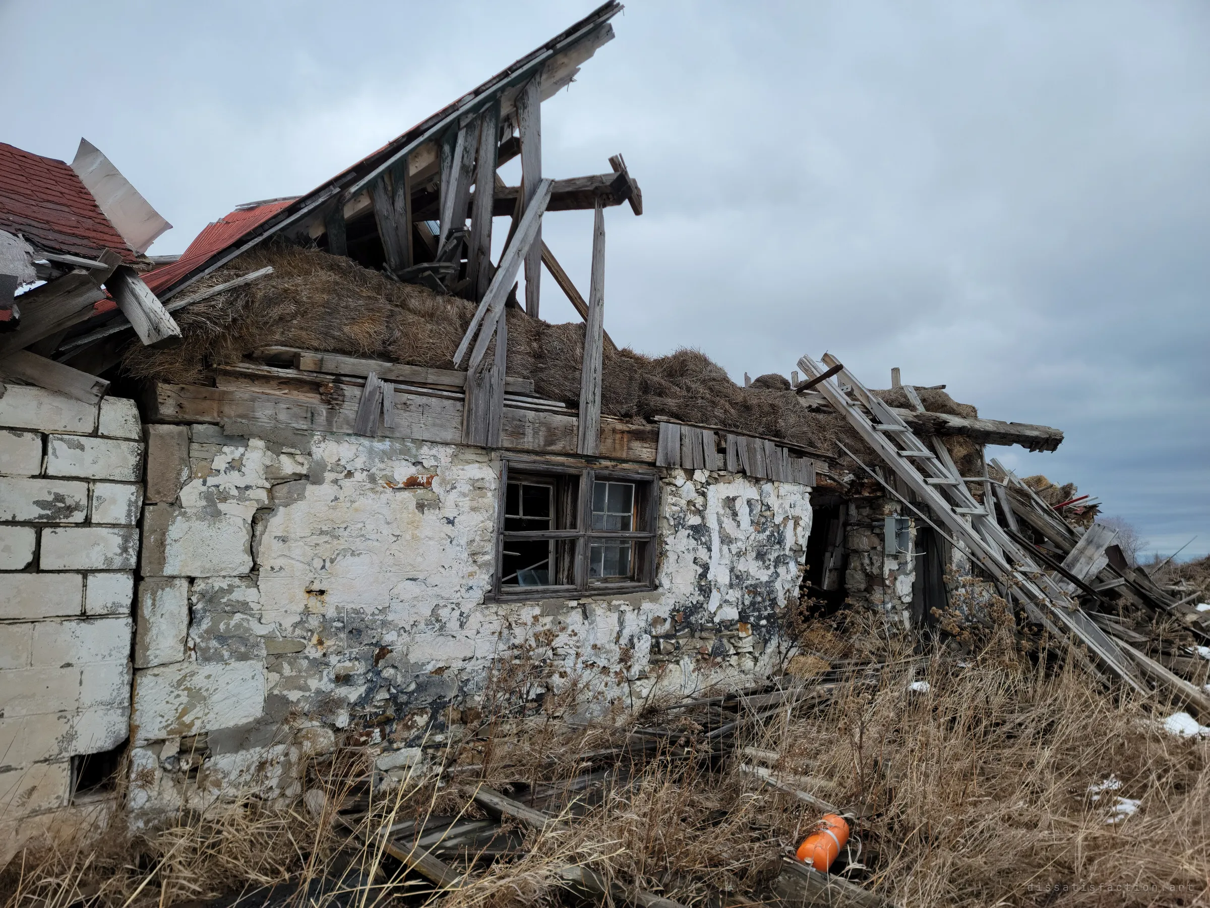 The stone wall of the collapsed barn, flaking whitewash and an old multi-pane window below a roof of exposed rafters and hay, with a ladder leaning into the wreckage. An orange tank lies in the grass.
