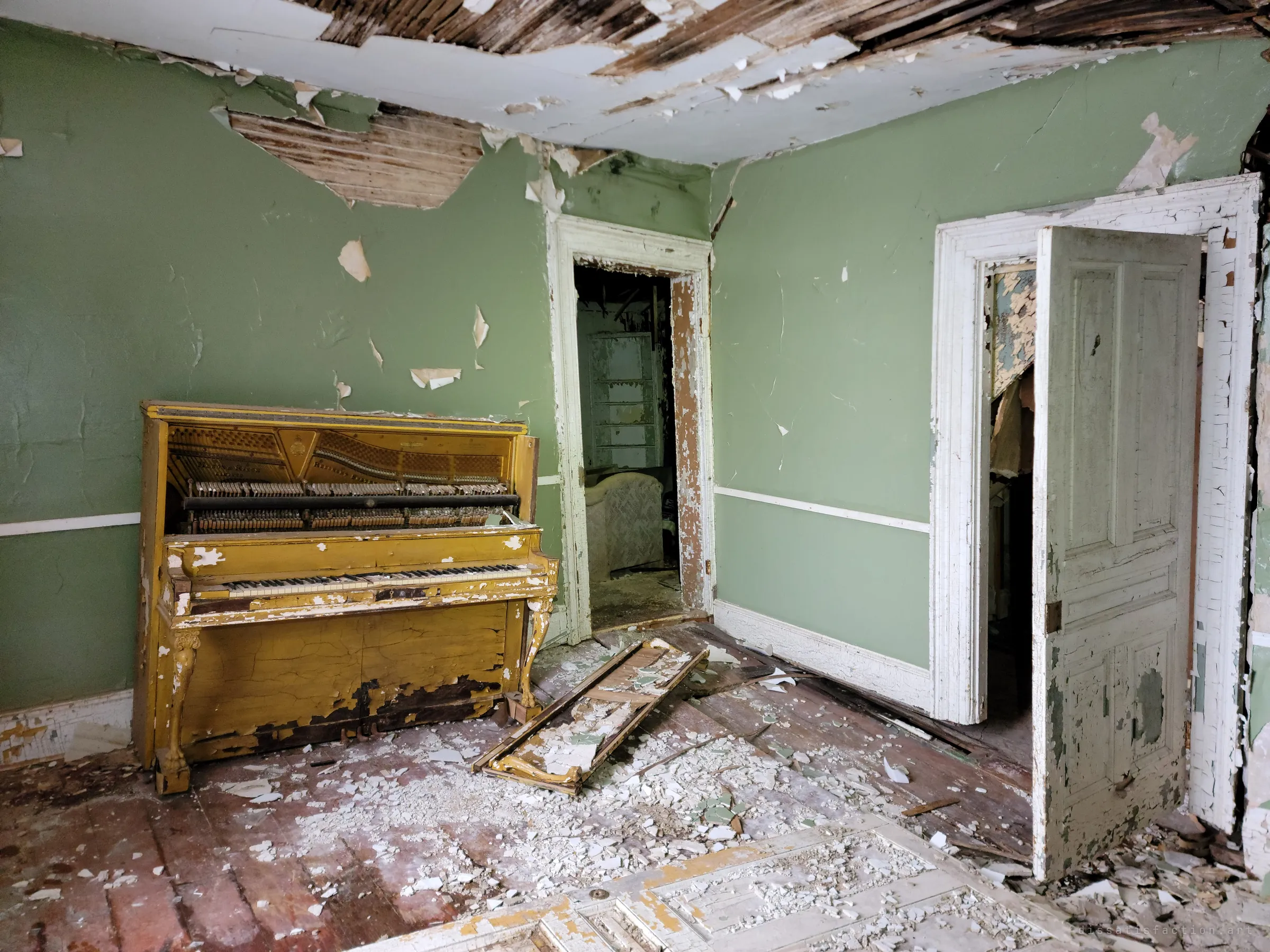 An upright piano with peeling yellow paint stands in a green room, its front panel off and plaster from the ceiling scattered across the floor. It is the centrepiece of the house's interior.