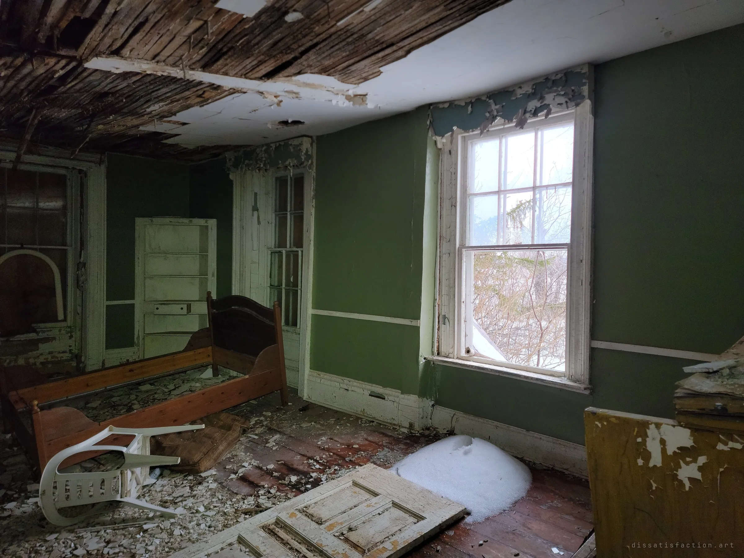 A bedroom with a wooden bed frame, an overturned plastic chair and a mound of snow blown in under the window, the ceiling stripped back to lath. Only trees show through the window.