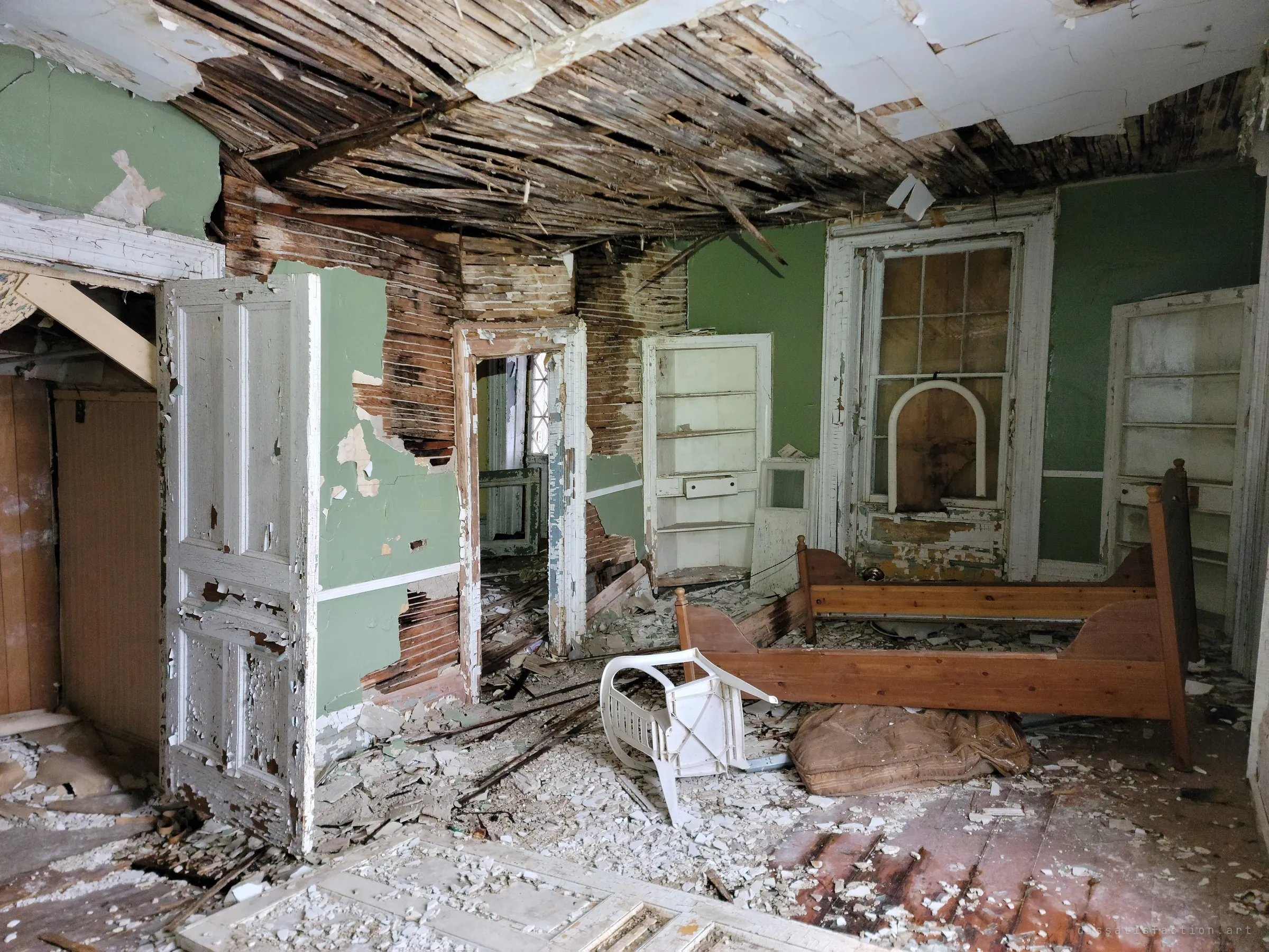 The same bedroom seen from the doorway, lath ceiling hanging down over built-in shelves, the bed frame and a toppled chair. Doorways lead into further ruined rooms.