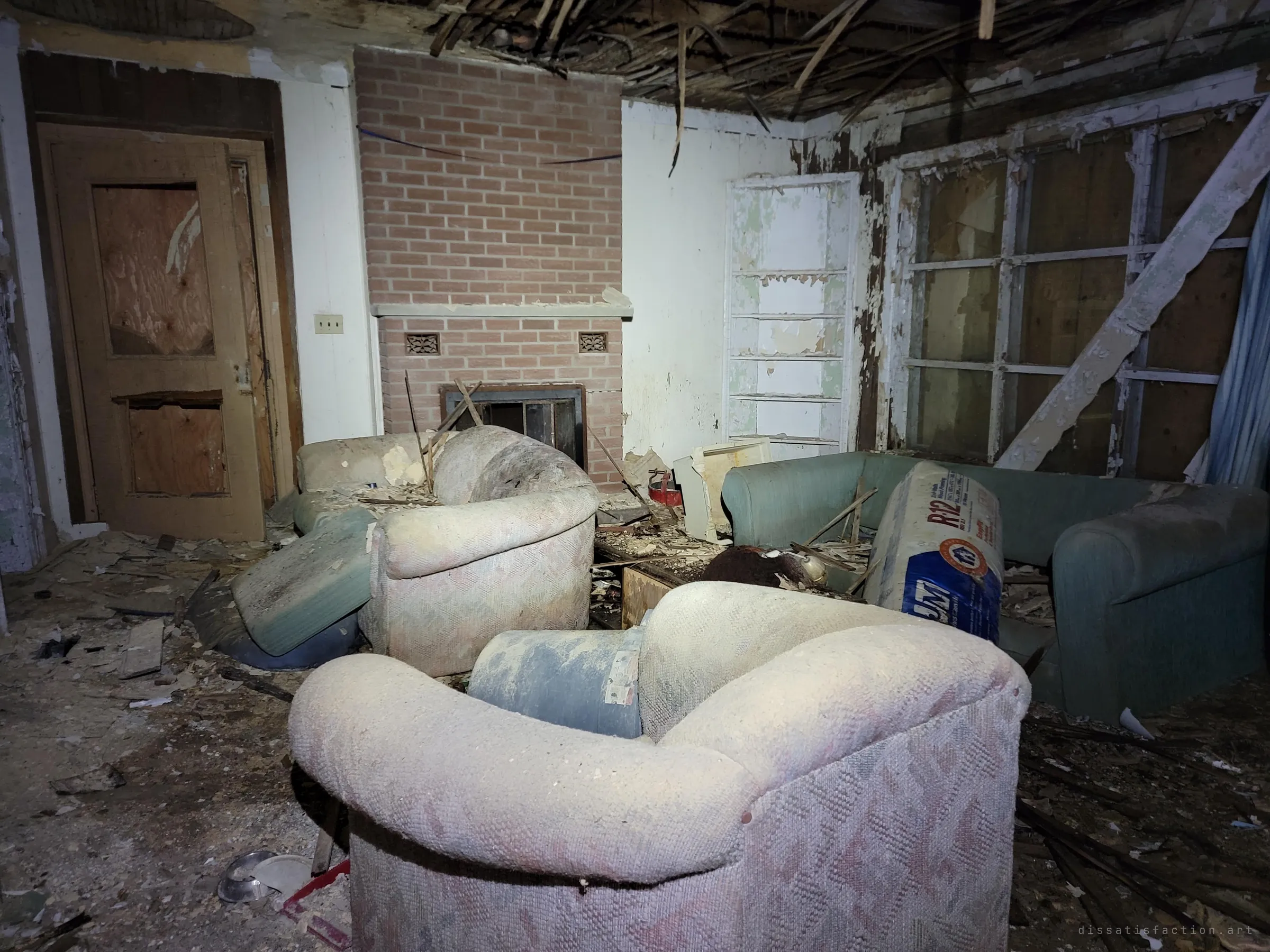 A living room buried in fallen ceiling lath, with dusty armchairs and a sofa gathered around a brick fireplace. The furniture is still arranged as if someone might sit down again.