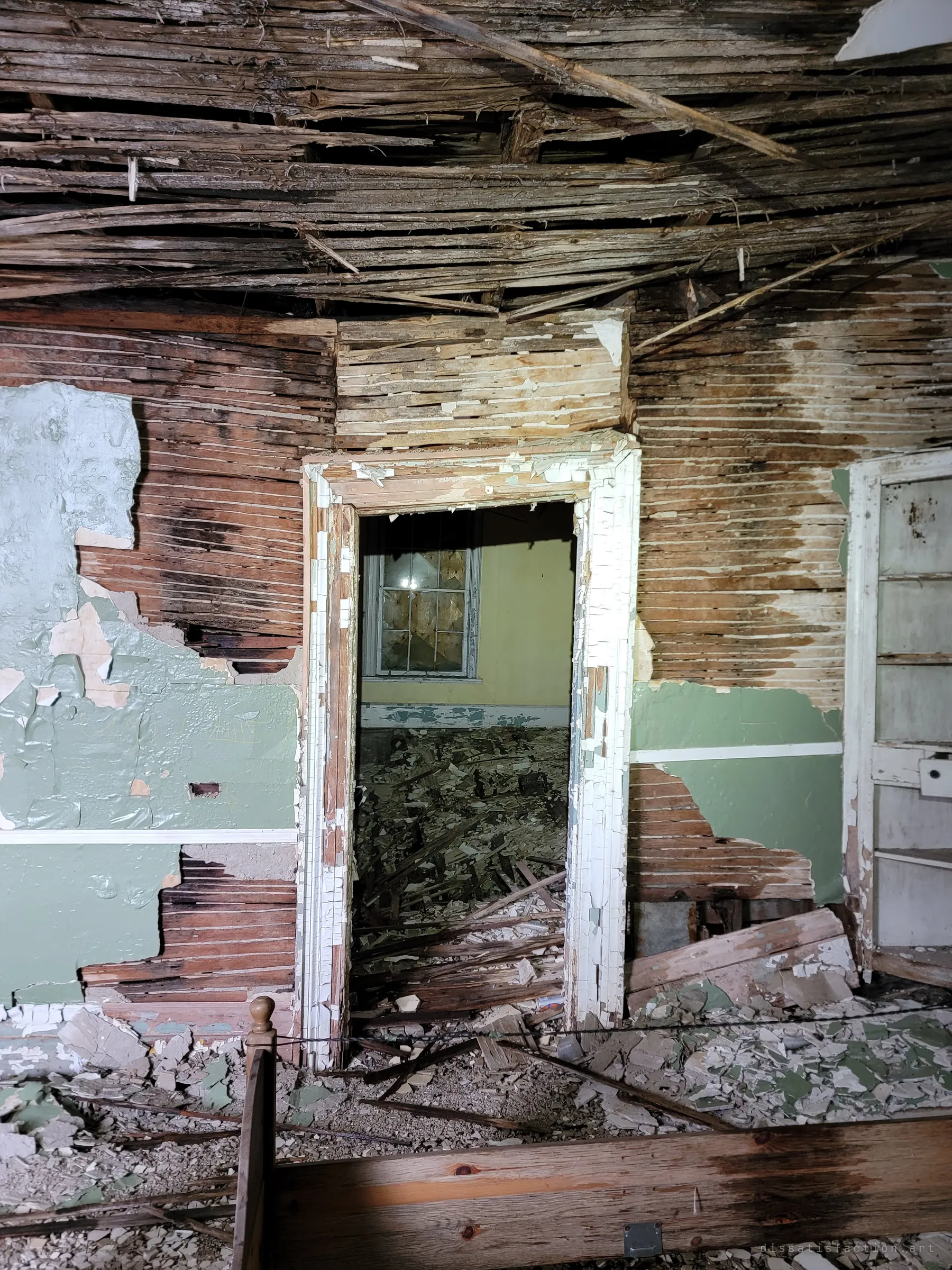 Plaster has fallen away to bare wooden lath, framing a doorway into a yellow room with a boarded window and a floor of debris. A rotting bed frame lies in the foreground.