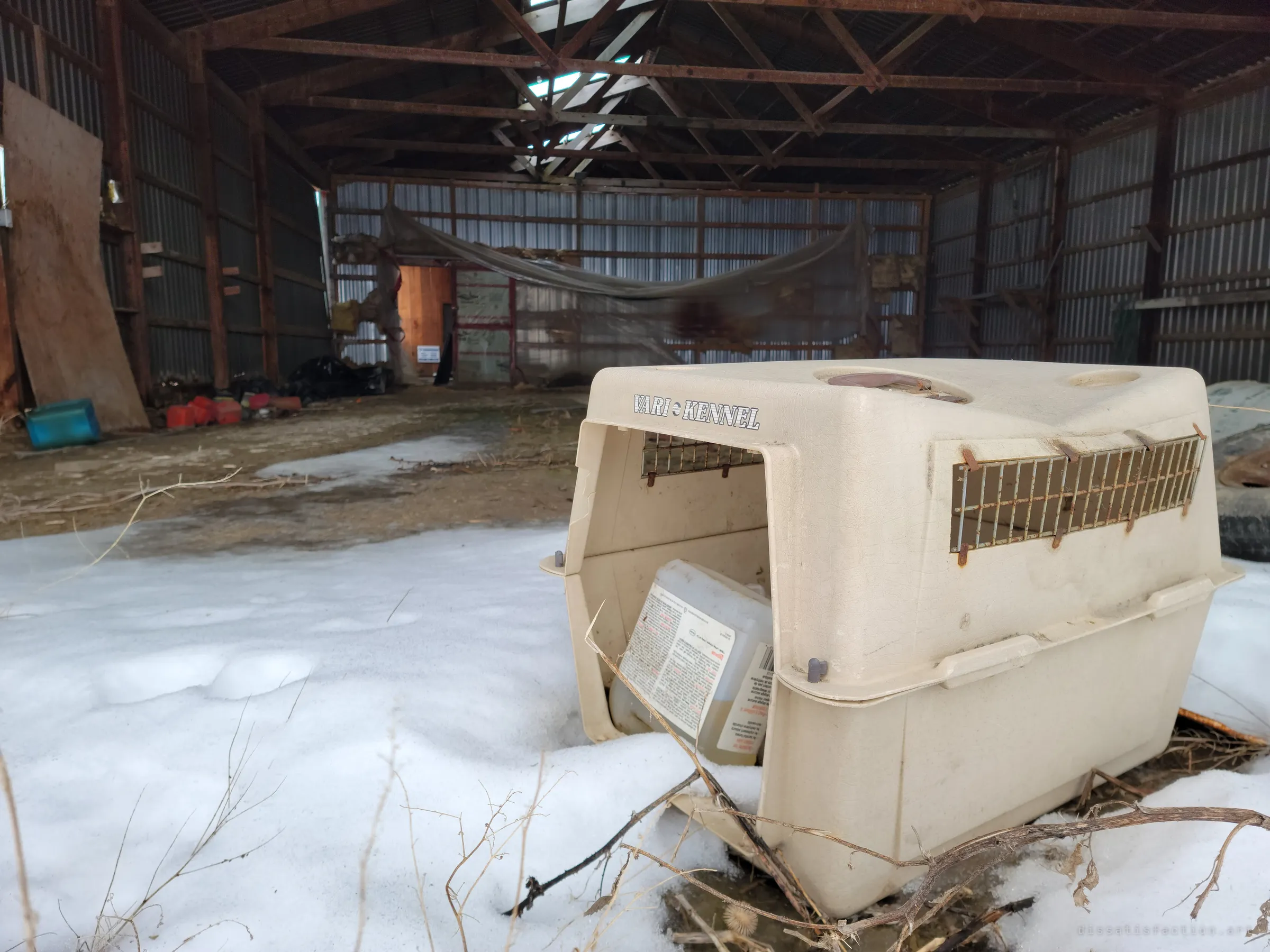 An empty pet carrier sits in the snow at the mouth of a metal pole barn, with torn plastic sheeting hanging across the back wall. Light comes through gaps in the roof.