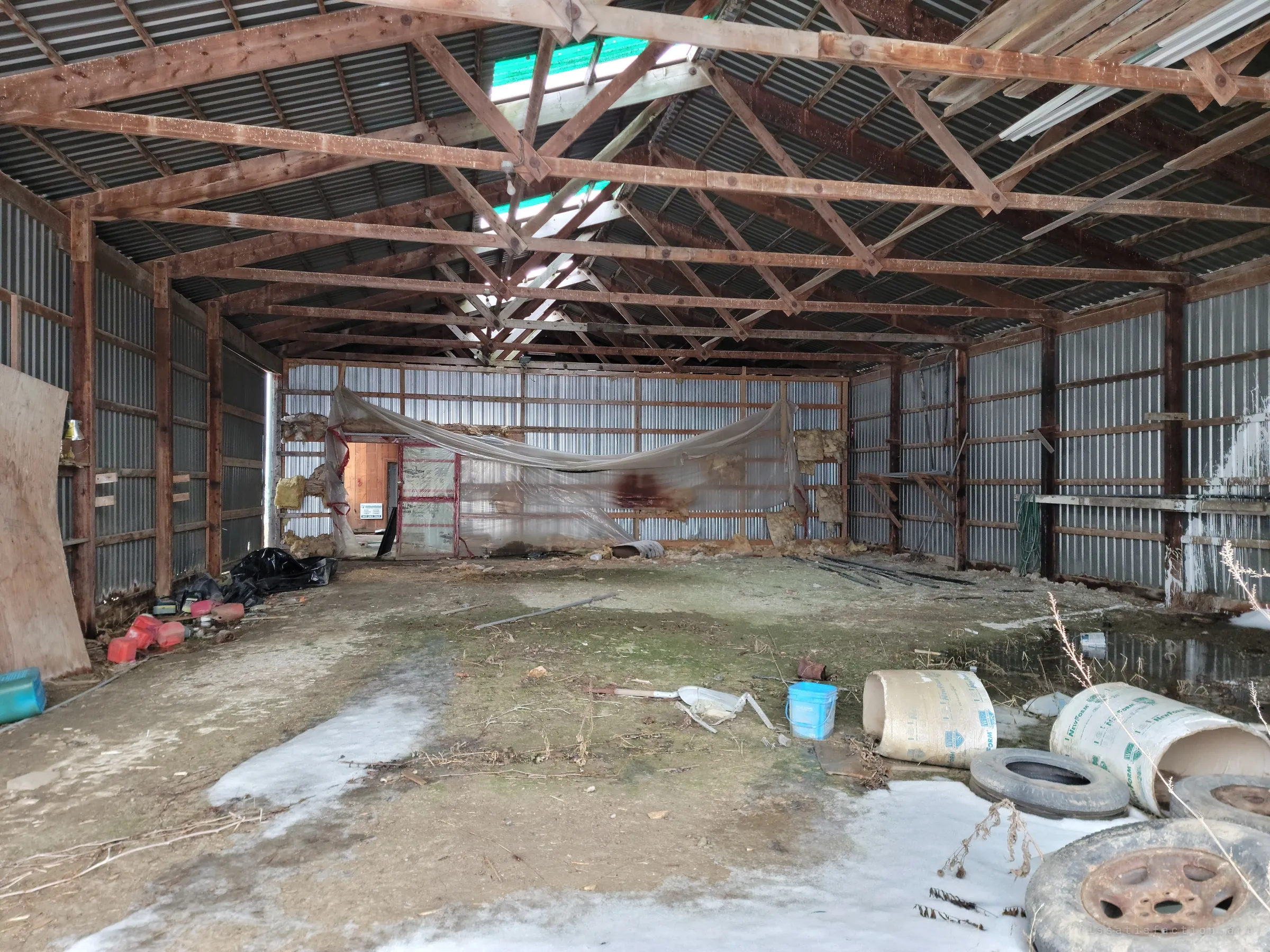 The full length of the pole barn under its wooden trusses, with a sagging plastic curtain, old tires and foam tubes scattered on a wet, icy floor.