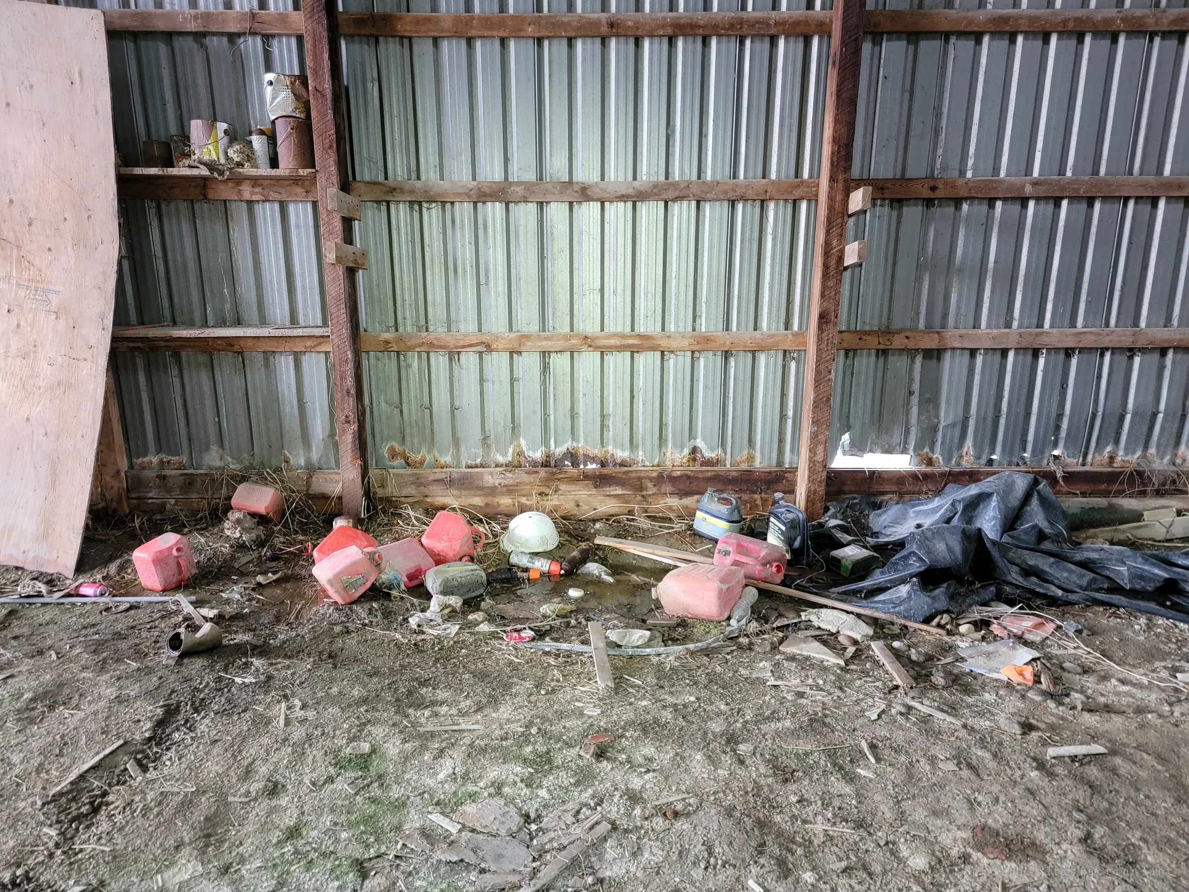 Red fuel cans, oil jugs and a hard hat lie in a puddle along the barn's corrugated wall, below a shelf of rusted paint tins.