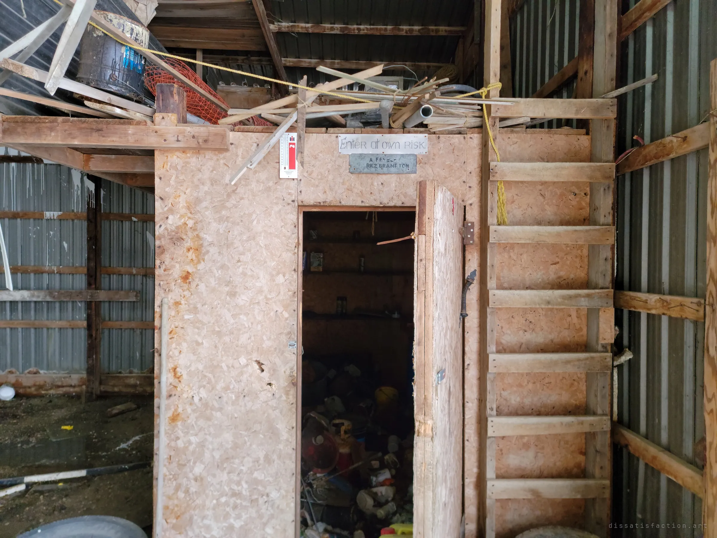 A chipboard workshop inside the barn with a hand-lettered warning sign above its open door and a ladder climbing to a loft piled with scrap wood.