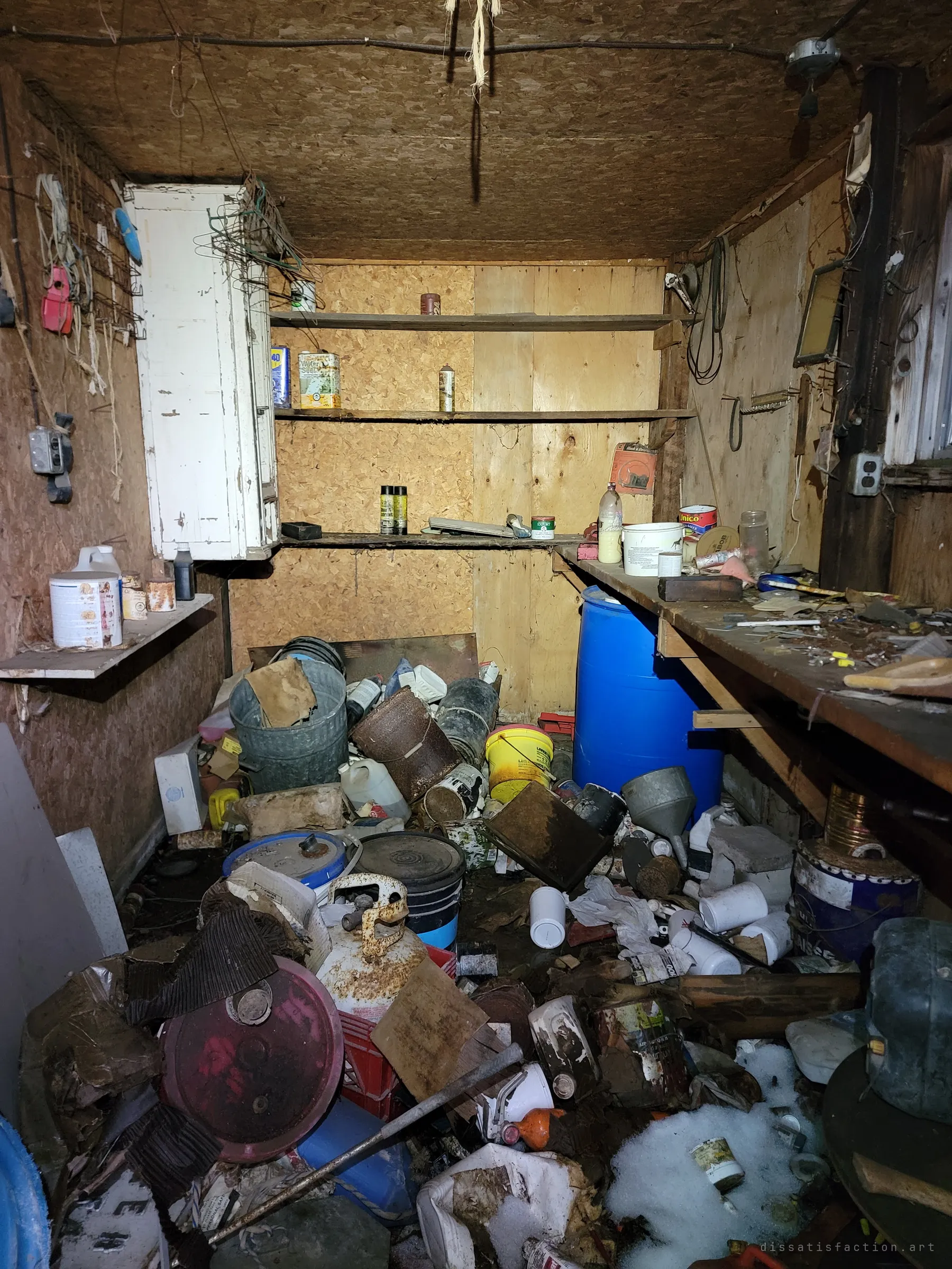 Inside the workshop: shelves of old cans and sprays above a floor heaped with buckets, drums, a propane tank and a blue barrel beside the workbench.