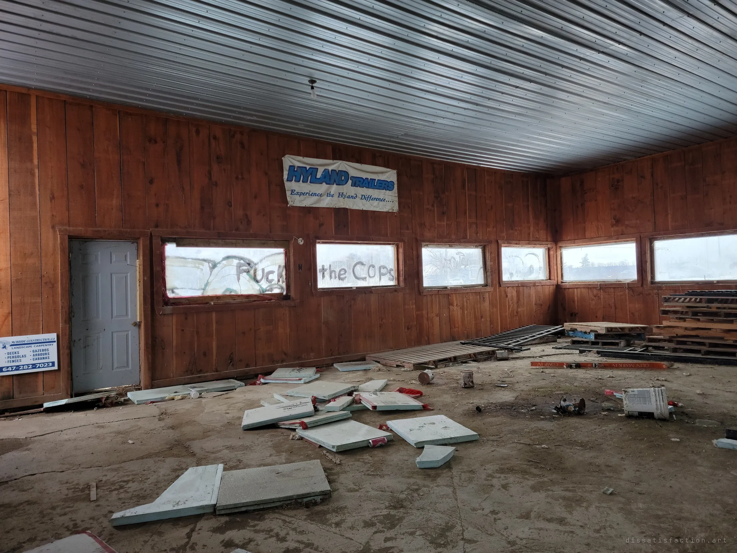 A wood-panelled showroom with a corrugated metal ceiling, its floor littered with broken foam insulation and pallets. Graffiti on the fogged windows and an old dealer banner show how the place turned from business to hangout.