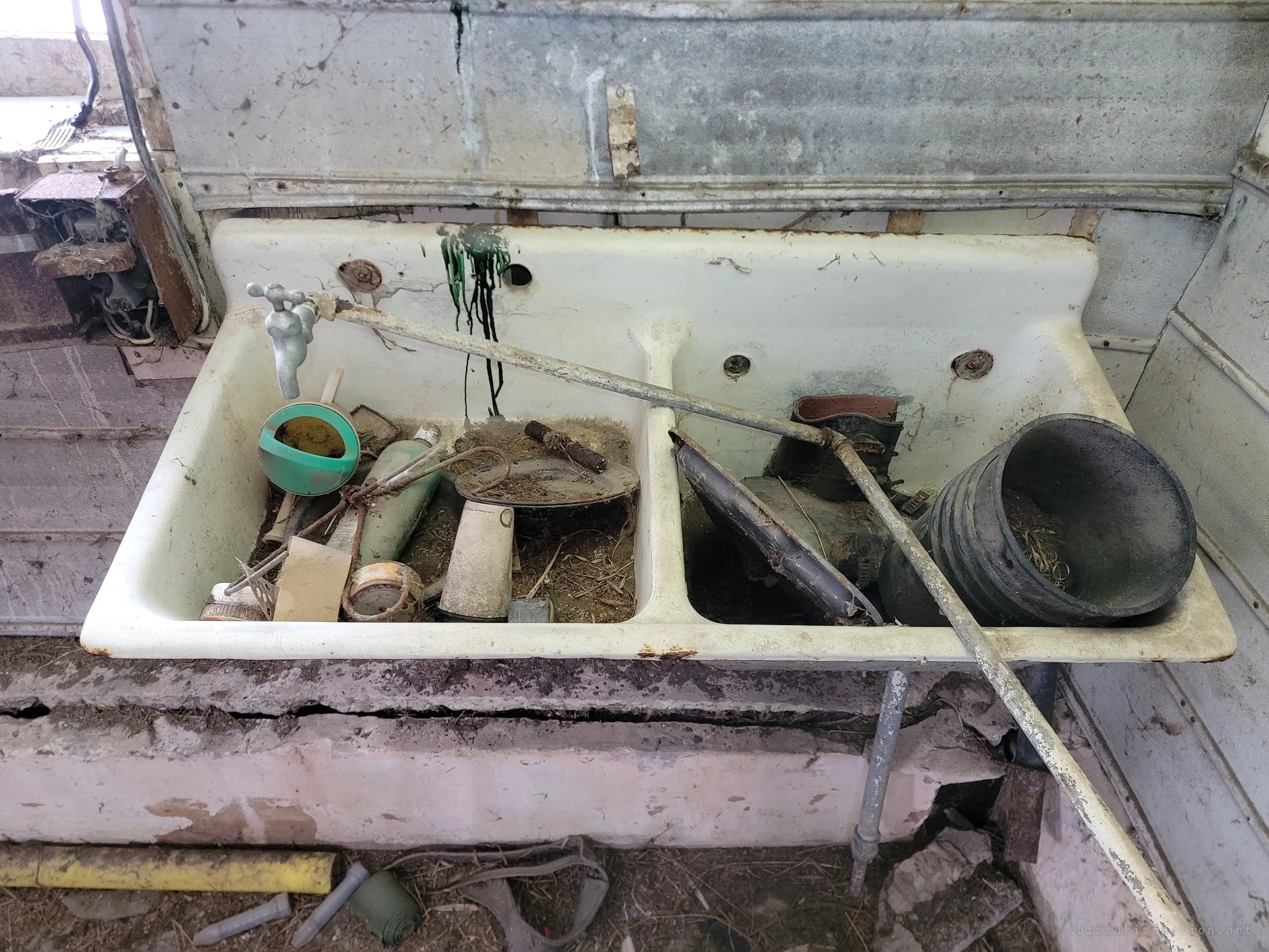 An old double-basin enamel farm sink crammed with rusted tools, a watering can, boots and a black bucket, under a galvanised backsplash. Decades of chores left where they were dropped.