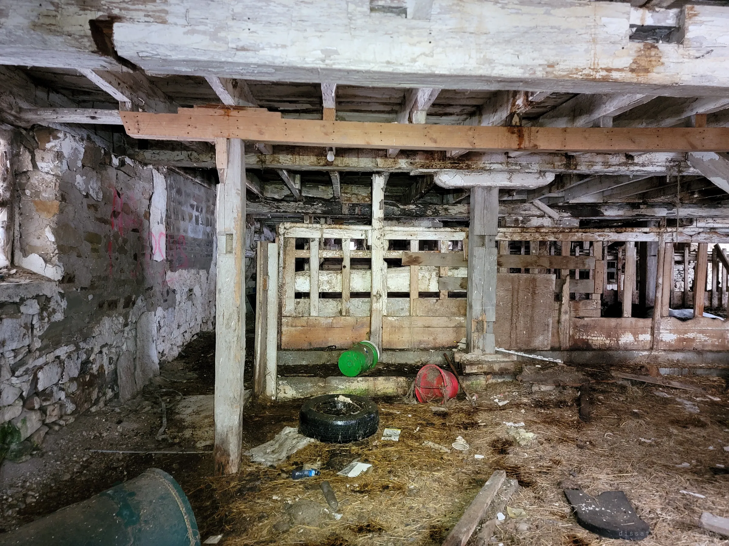 The low stable level of a barn: fieldstone walls, whitewashed beams and wooden stall gates, with a tyre and plastic buckets in the straw. The heavy timber frame still holds up the floor above.