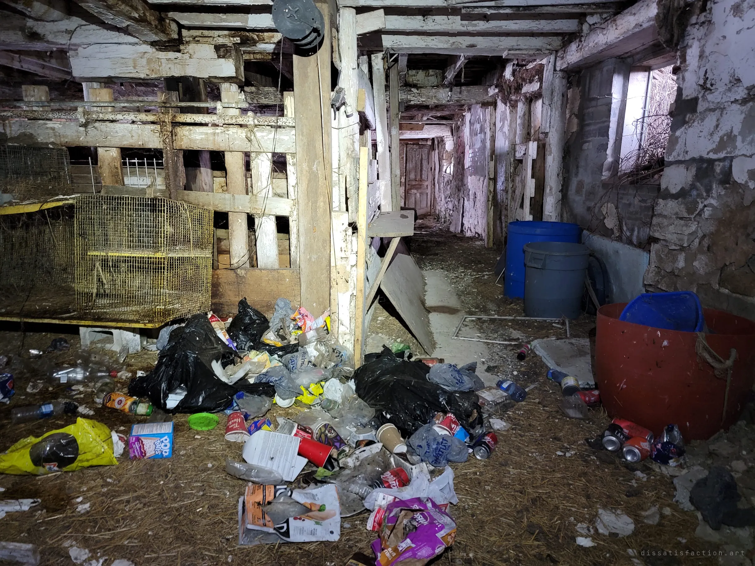 A long whitewashed stable corridor strewn with garbage bags, cans and snack wrappers beside old animal cages and plastic drums. Recent visitors have left their mark on the old barn.