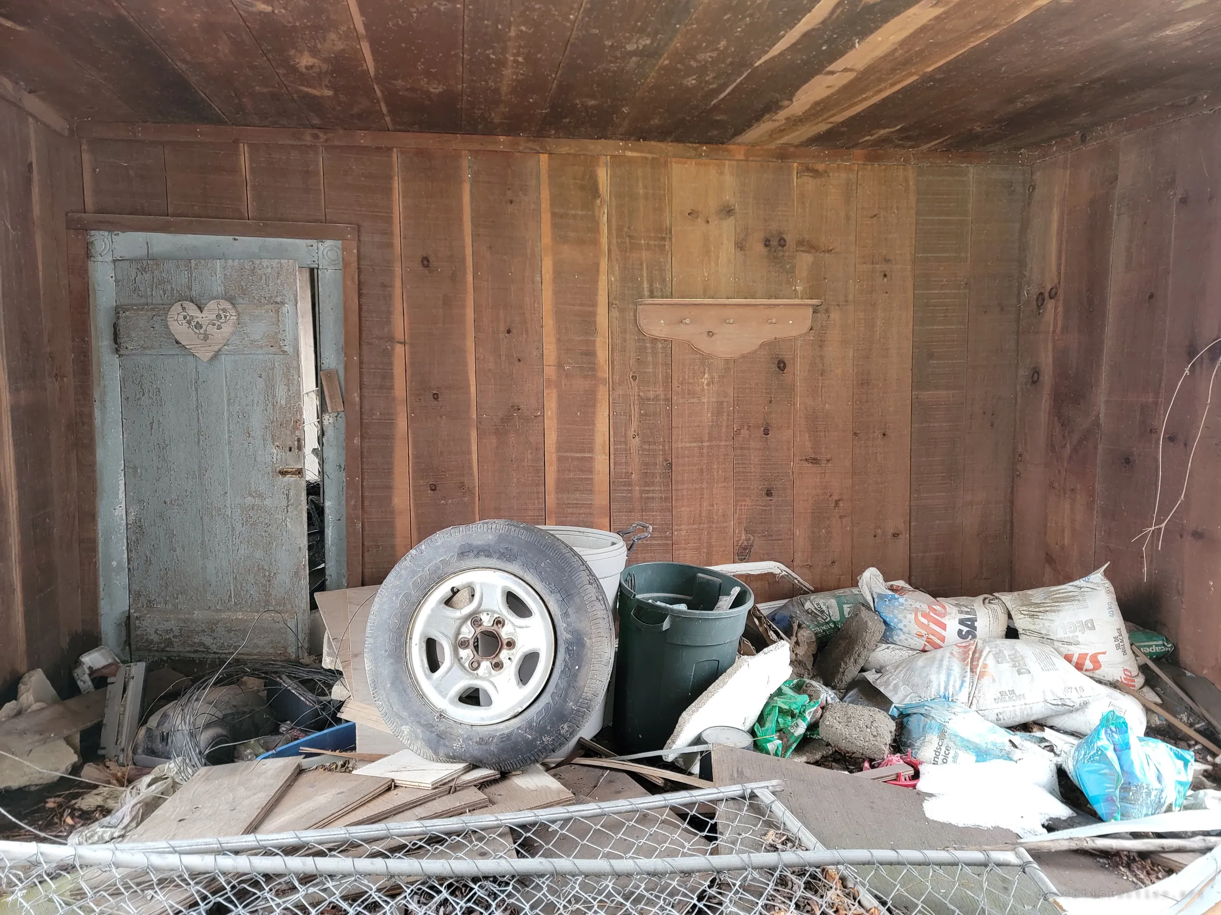 A plank-walled room with a pale blue door decorated with a wooden heart, a pile of tyres, bins and split salt bags heaped in front of it. A homely touch left among the junk.