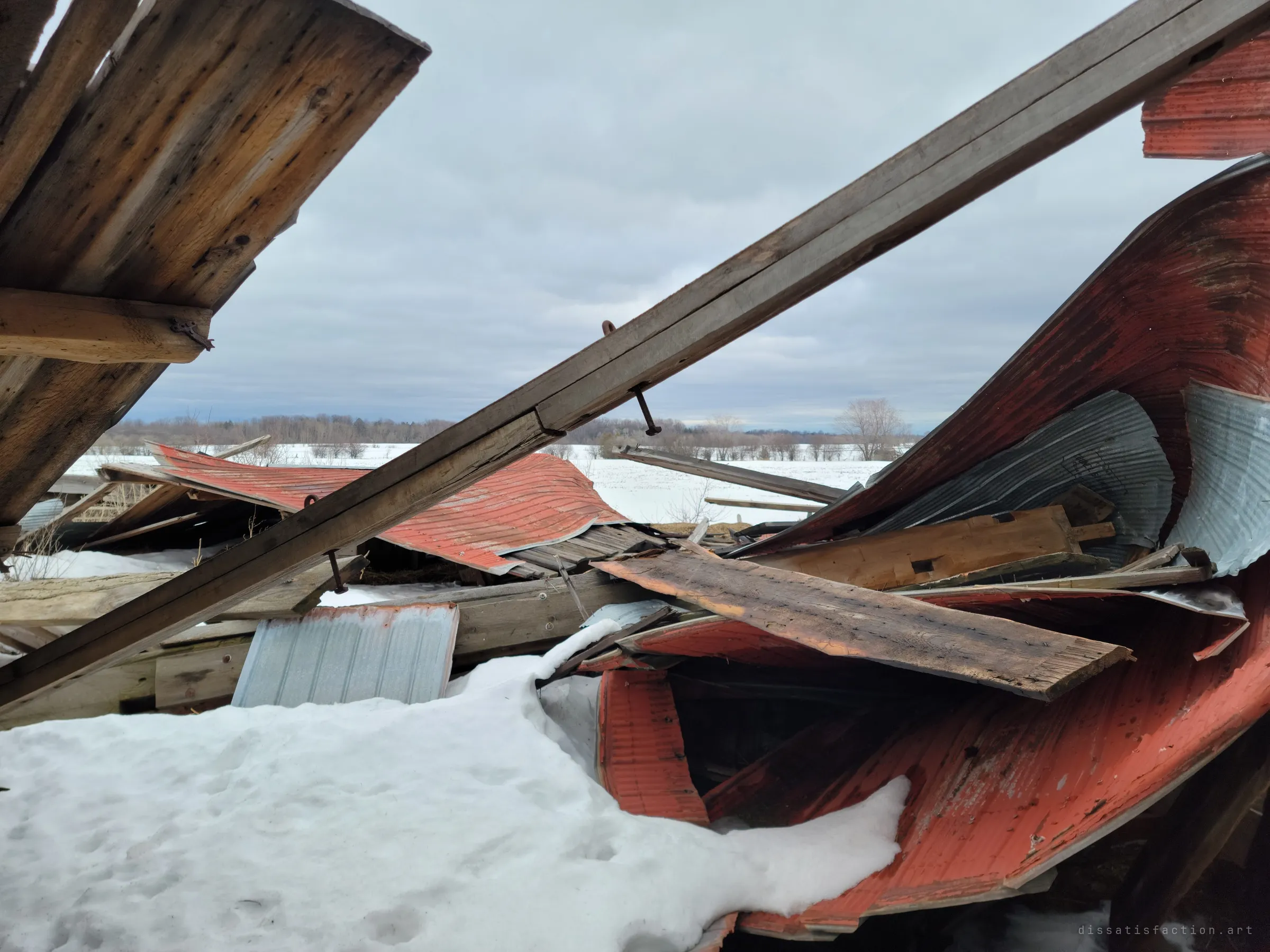 The wreck of a collapsed barn, red metal roofing twisted around broken beams, half buried in snow. Flat snowy fields and a tree line stretch out under a grey sky.