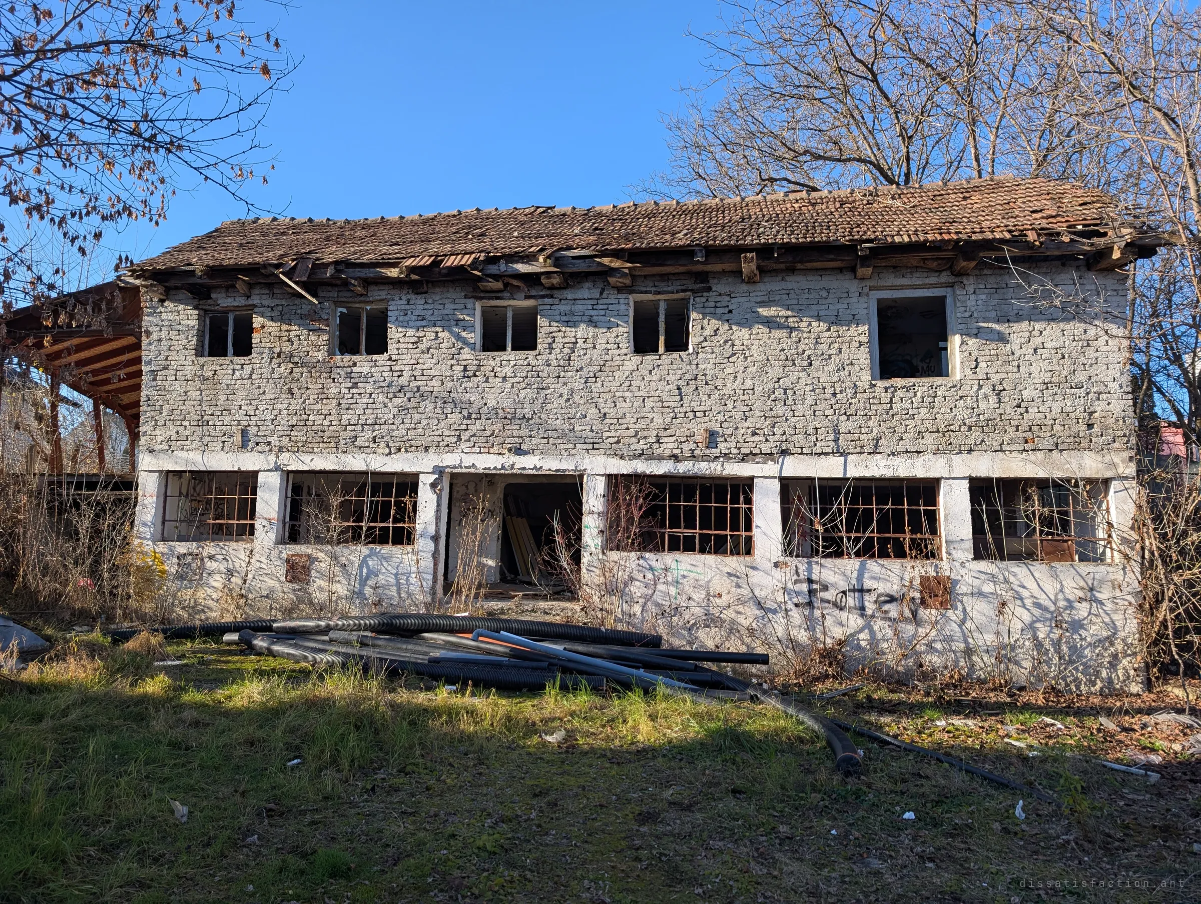 The whitewashed brick building from outside, its tiled roof caving in over empty upper windows and rusted bars below, with plastic pipes dumped in the grass. It sets the scene: a small forgotten block left to the weeds.
