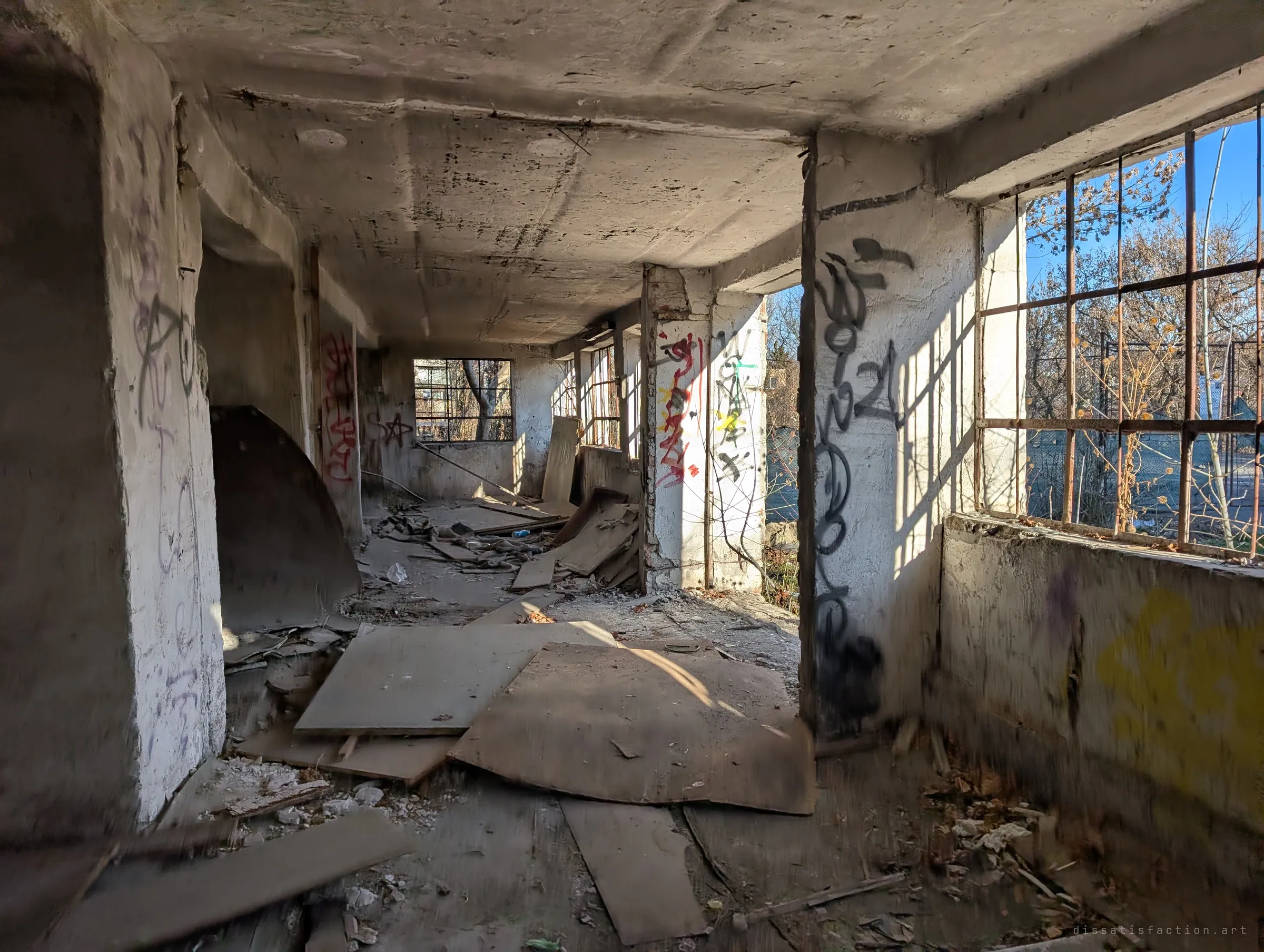 A ground-floor room of fallen boards and graffiti-covered pillars, low sun pouring through barred windows. Light and debris show how open the building now is to the outside.