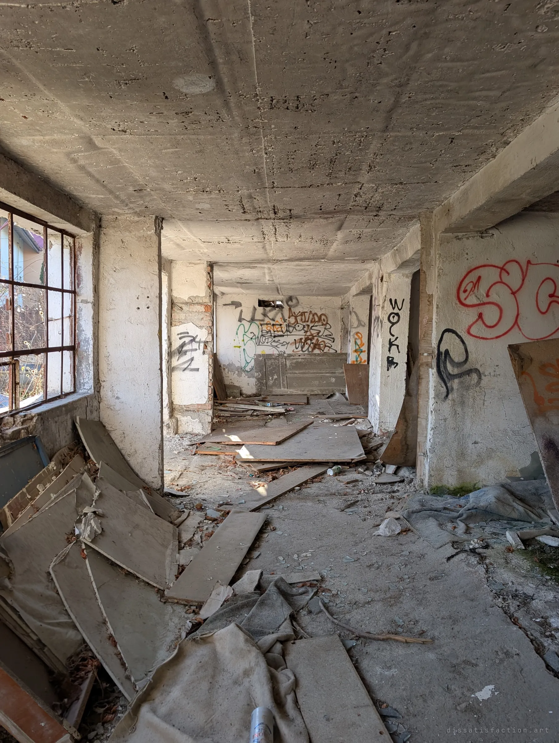 A long concrete corridor strewn with boards and broken glass, tags covering the far walls. The rough ceiling and bare pillars show the building stripped to its shell.