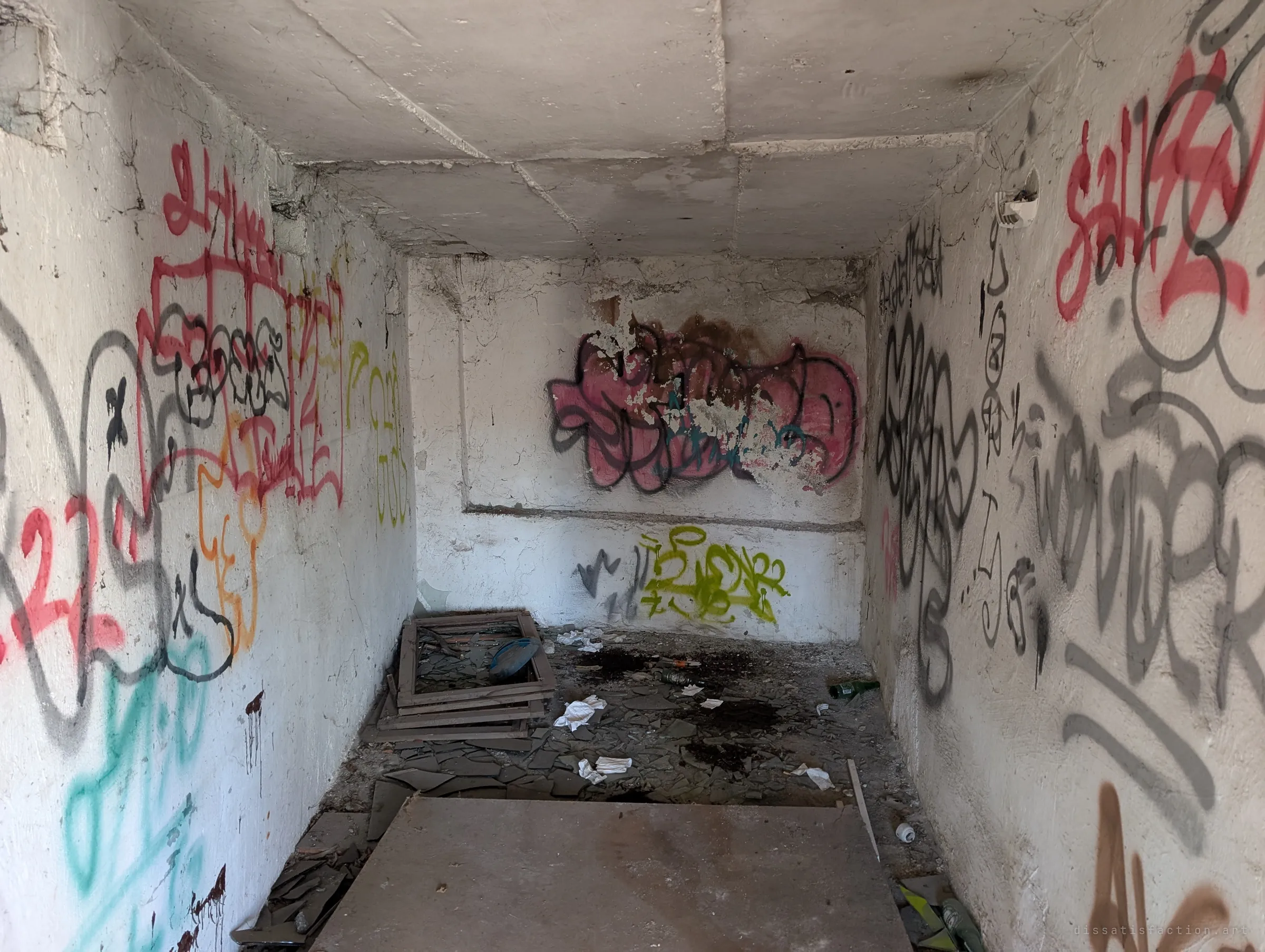 A small concrete room with every wall covered in layered graffiti, broken glass and window frames on the floor. It shows how thoroughly the place has been claimed by taggers.