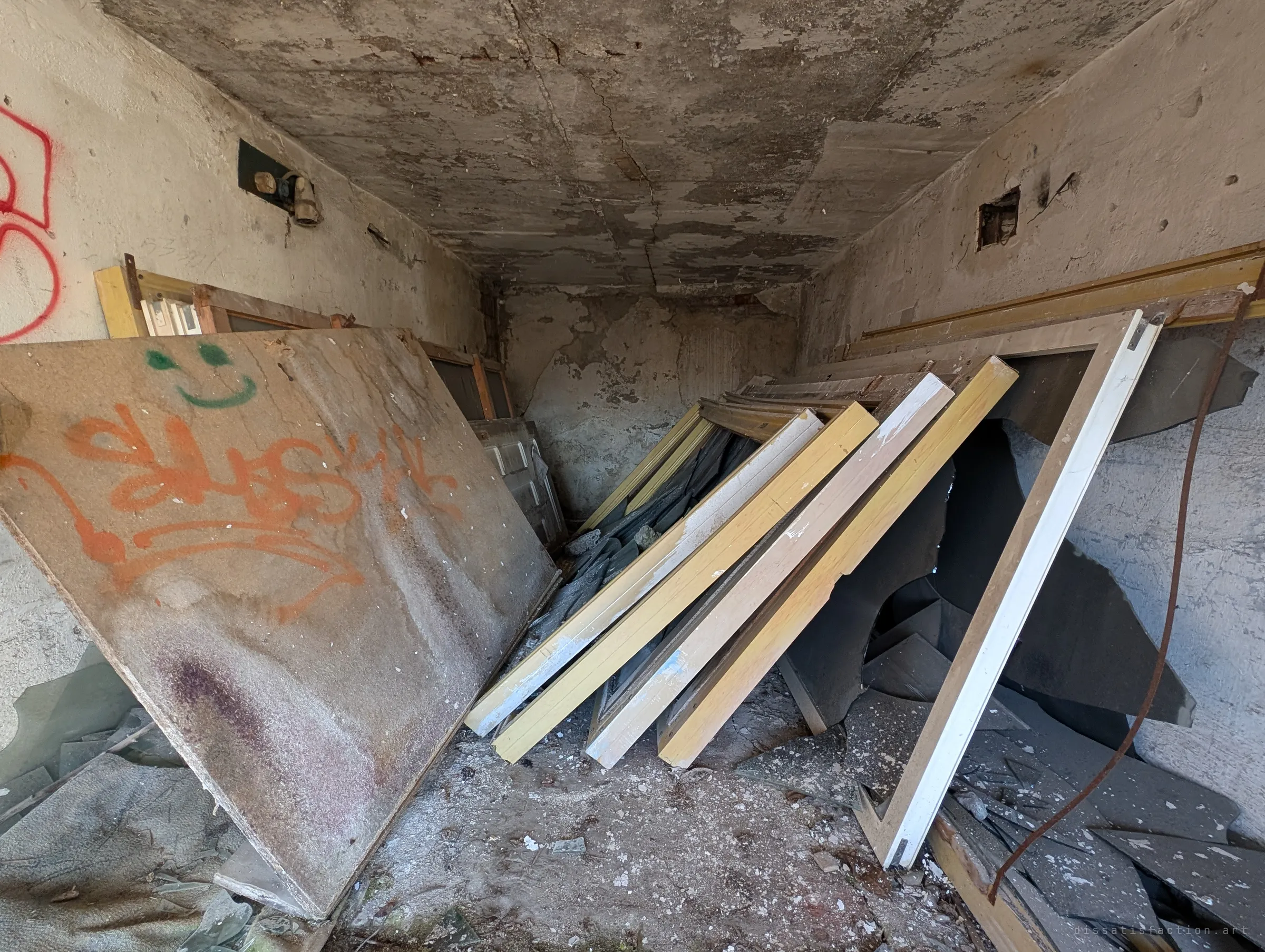 Old window frames and doors stacked against the walls of a peeling concrete room, a tagged board with a smiley leaning in front. Leftover fittings hint at a building gutted piece by piece.