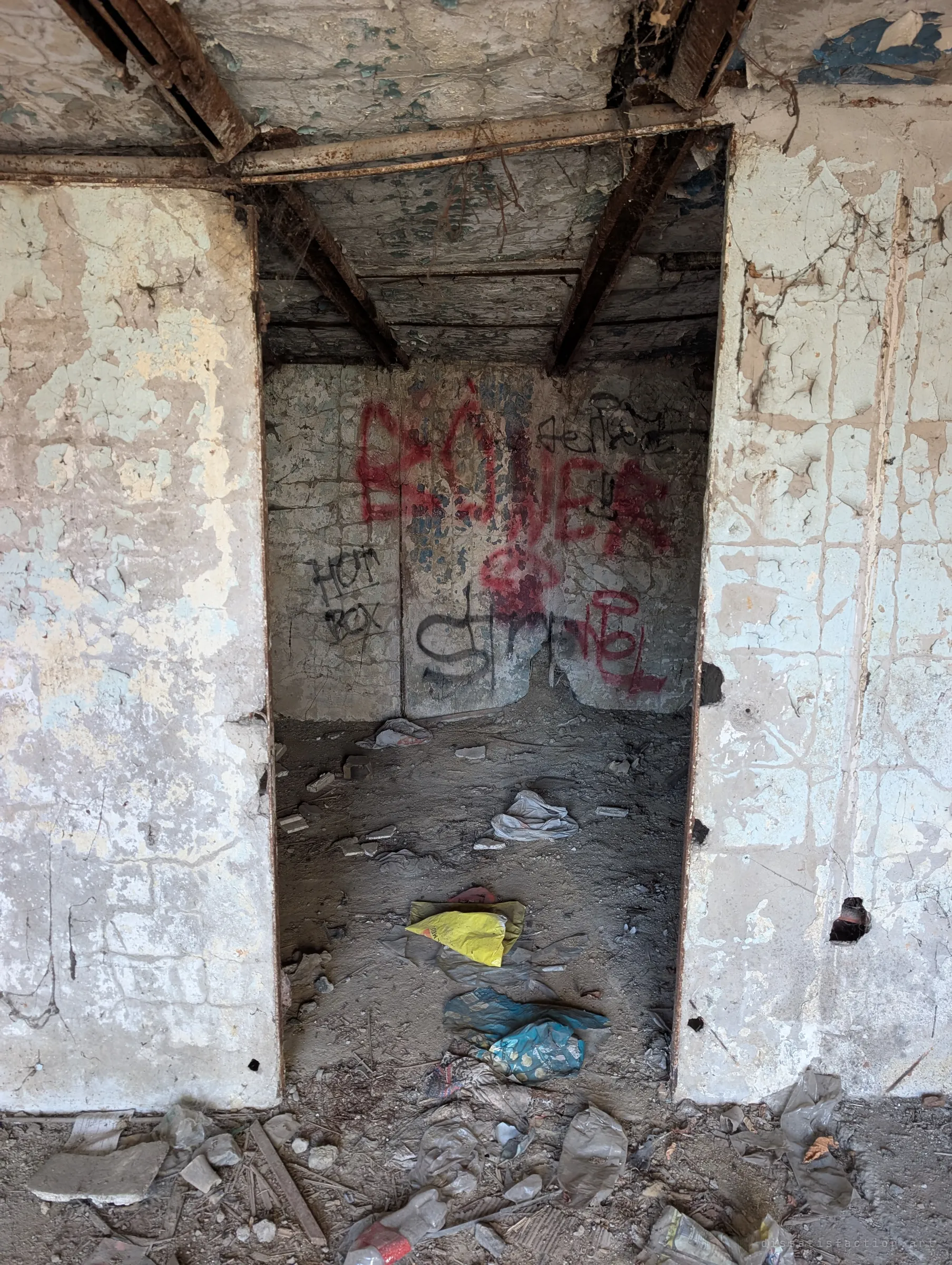 A narrow doorway under rusted steel beams opens into a small graffiti-marked room, plastic bags and rubble on the floor. The flaking paint and litter show years of neglect.