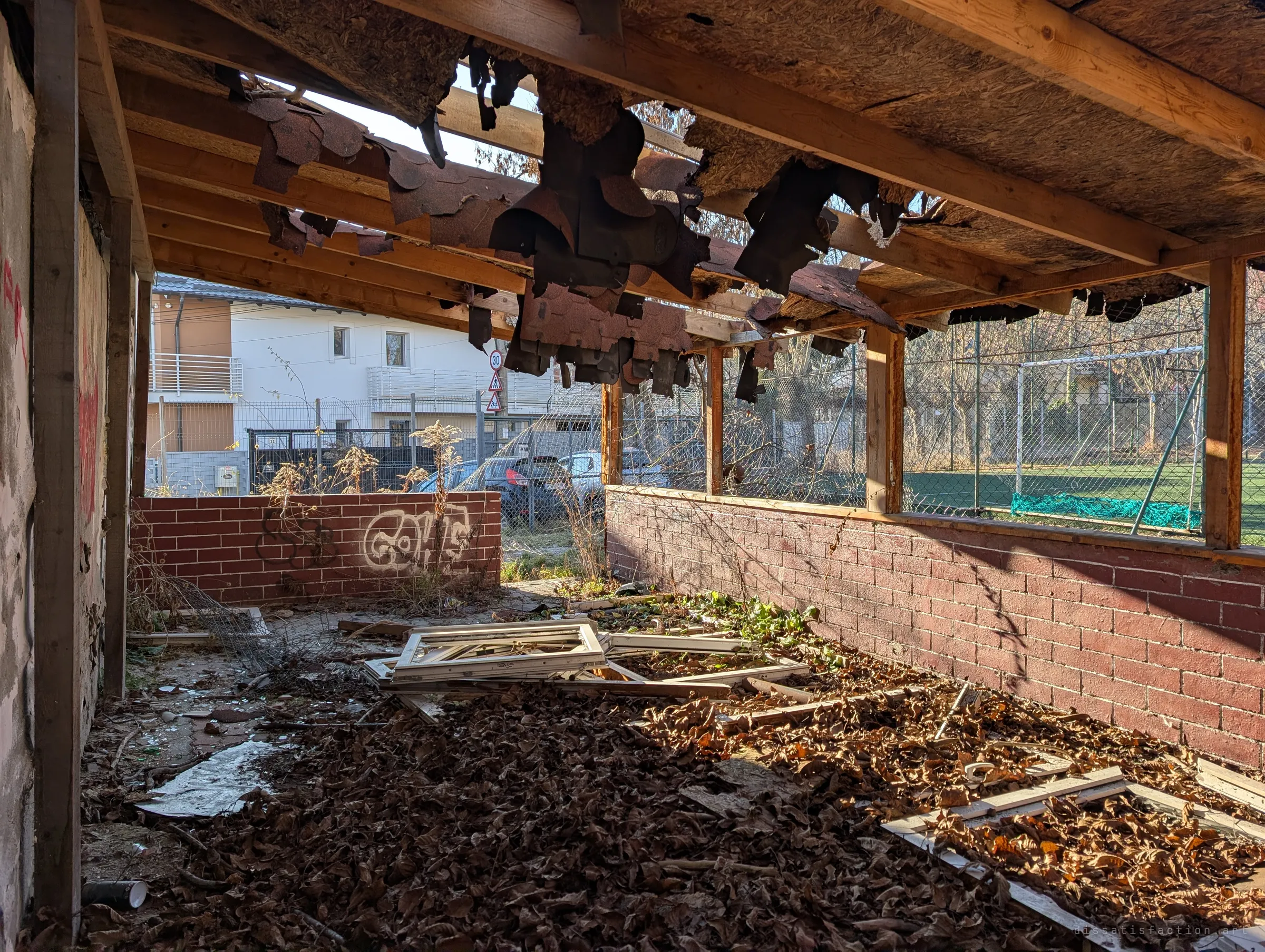 A roofless wooden pavilion carpeted in dry leaves and broken window frames, tattered roofing hanging overhead. Beyond the low brick wall, the everyday street carries on beside the ruin.
