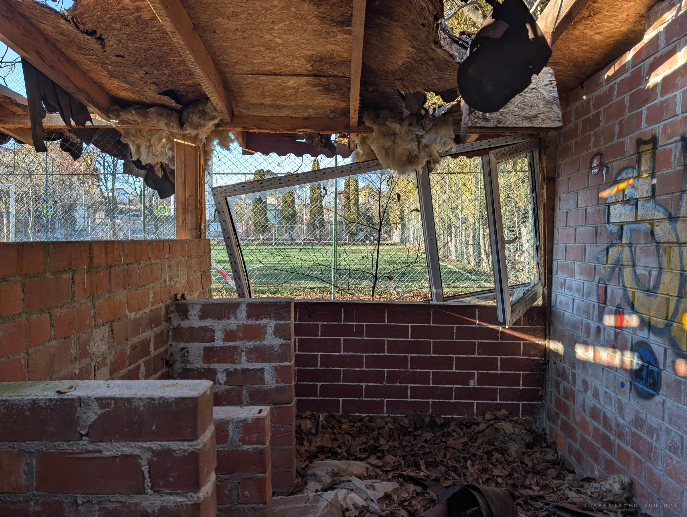 A corner of the pavilion with a PVC window hanging askew from its frame, insulation spilling from the rotten roof. Through it, a bright green pitch sits just past the fence.