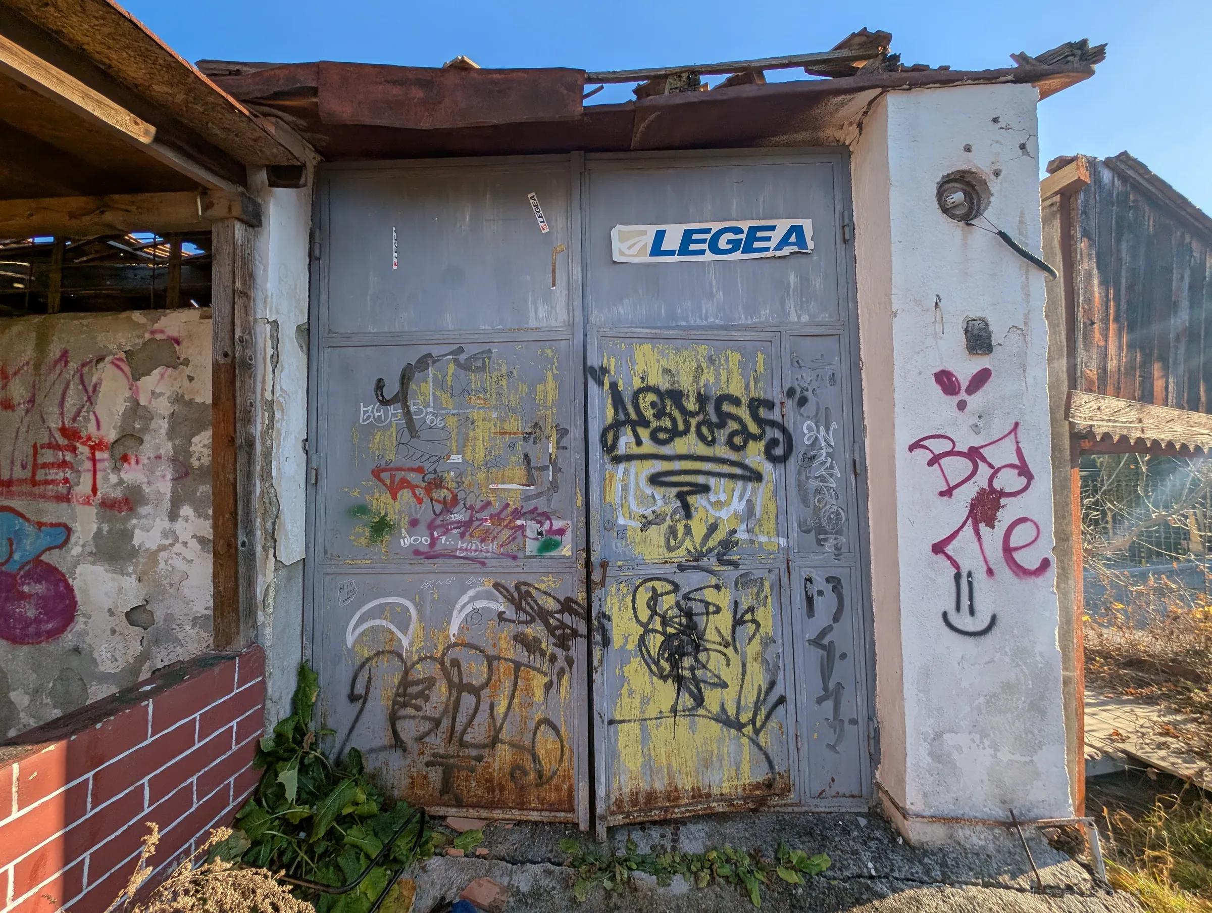 A pair of grey steel garage doors thick with tags and a sportswear sticker, under a sagging rusted roof edge. The doors are the building's sealed face, patched and painted over many times.