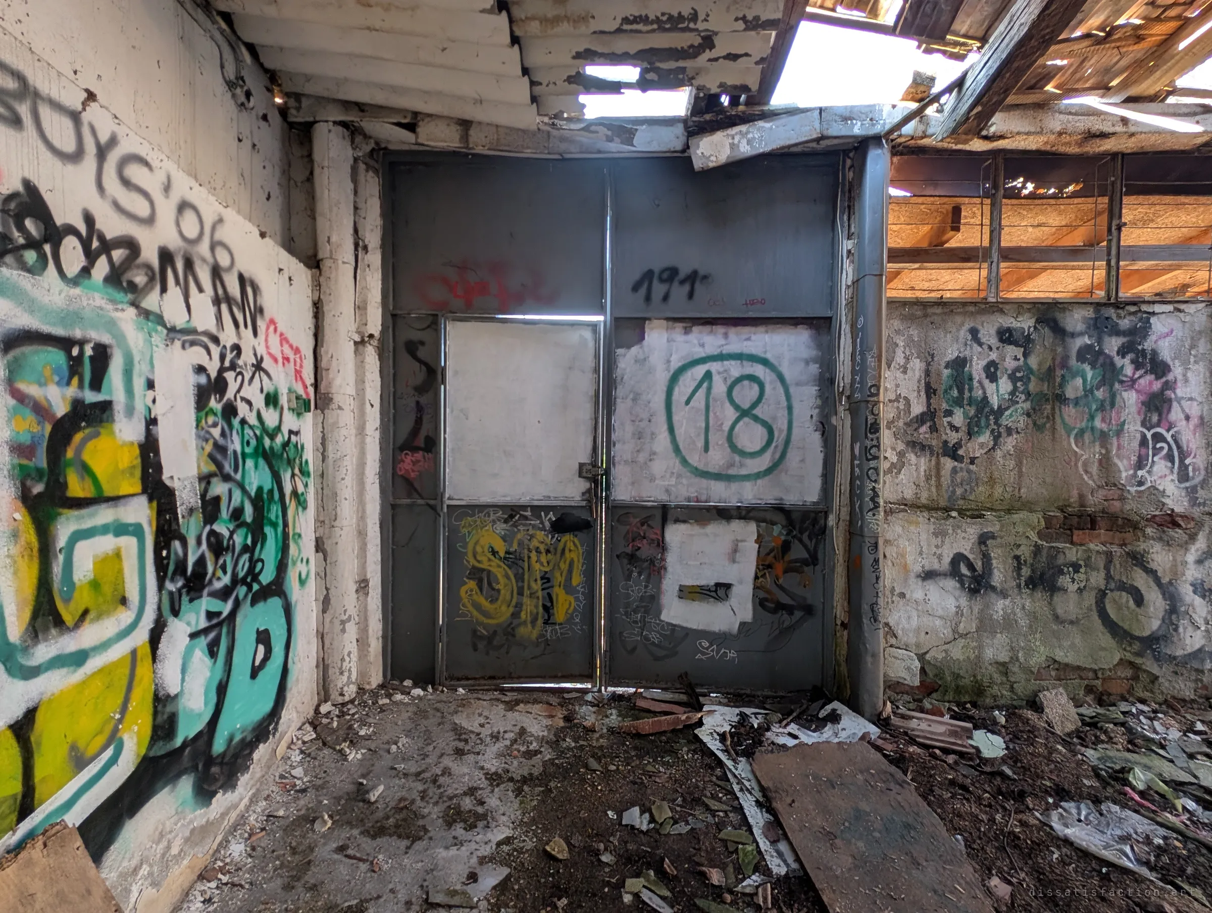 The inside of the steel doors, marked with a large painted number, beside walls buried under colourful graffiti and a roof open to the sky. It shows the garage bay abandoned and half exposed.