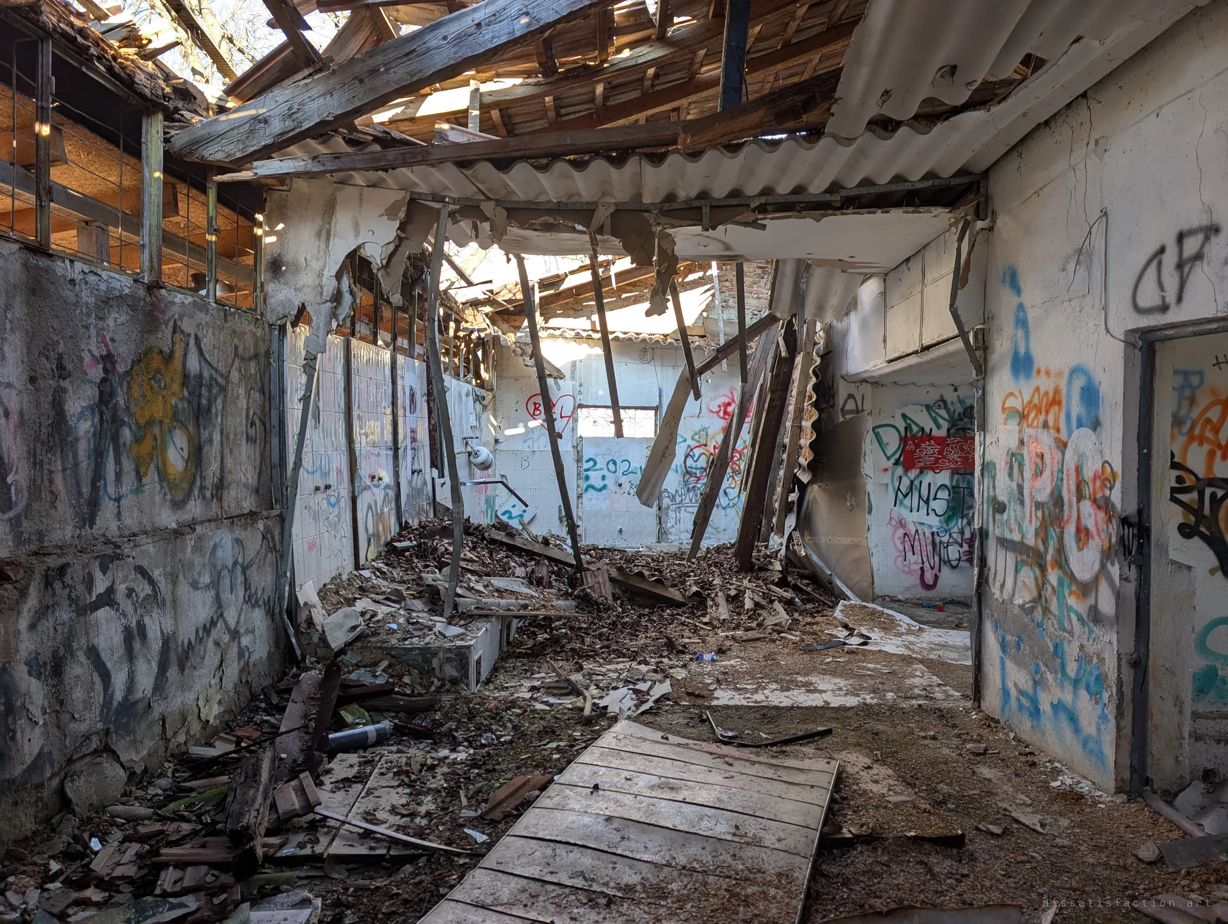 A long tiled washroom lies open to the sky, its roof collapsed into a heap of beams, sheeting and leaves between graffiti-covered walls. The row of wall fittings on the left hints at the shower stalls it once held.