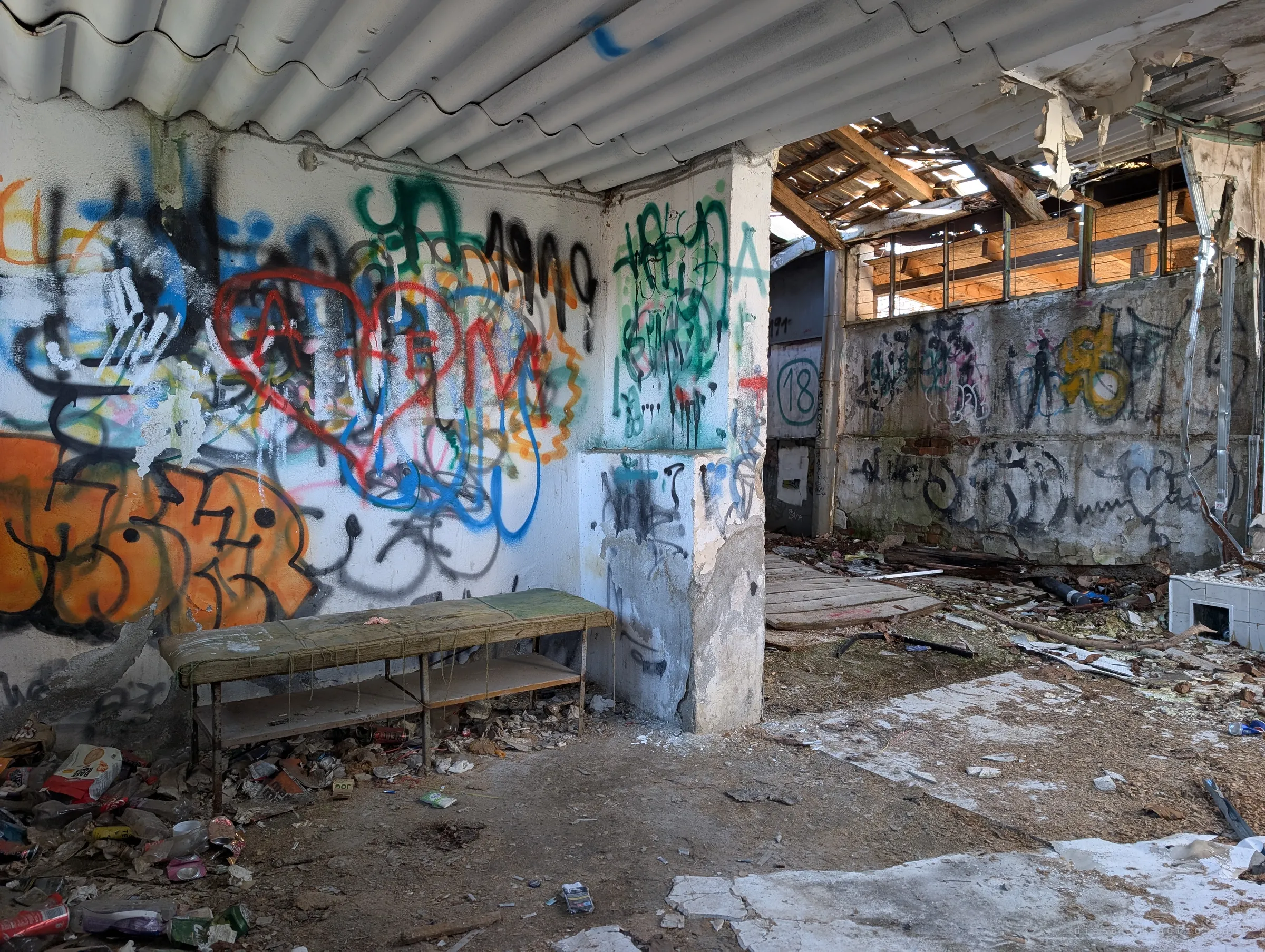 A padded bench with a mouldy green cover sits against a wall layered in graffiti, beside a painted cubicle number and a room whose roof has fallen in. Litter and cans pile up around its legs, the remains of later visitors.