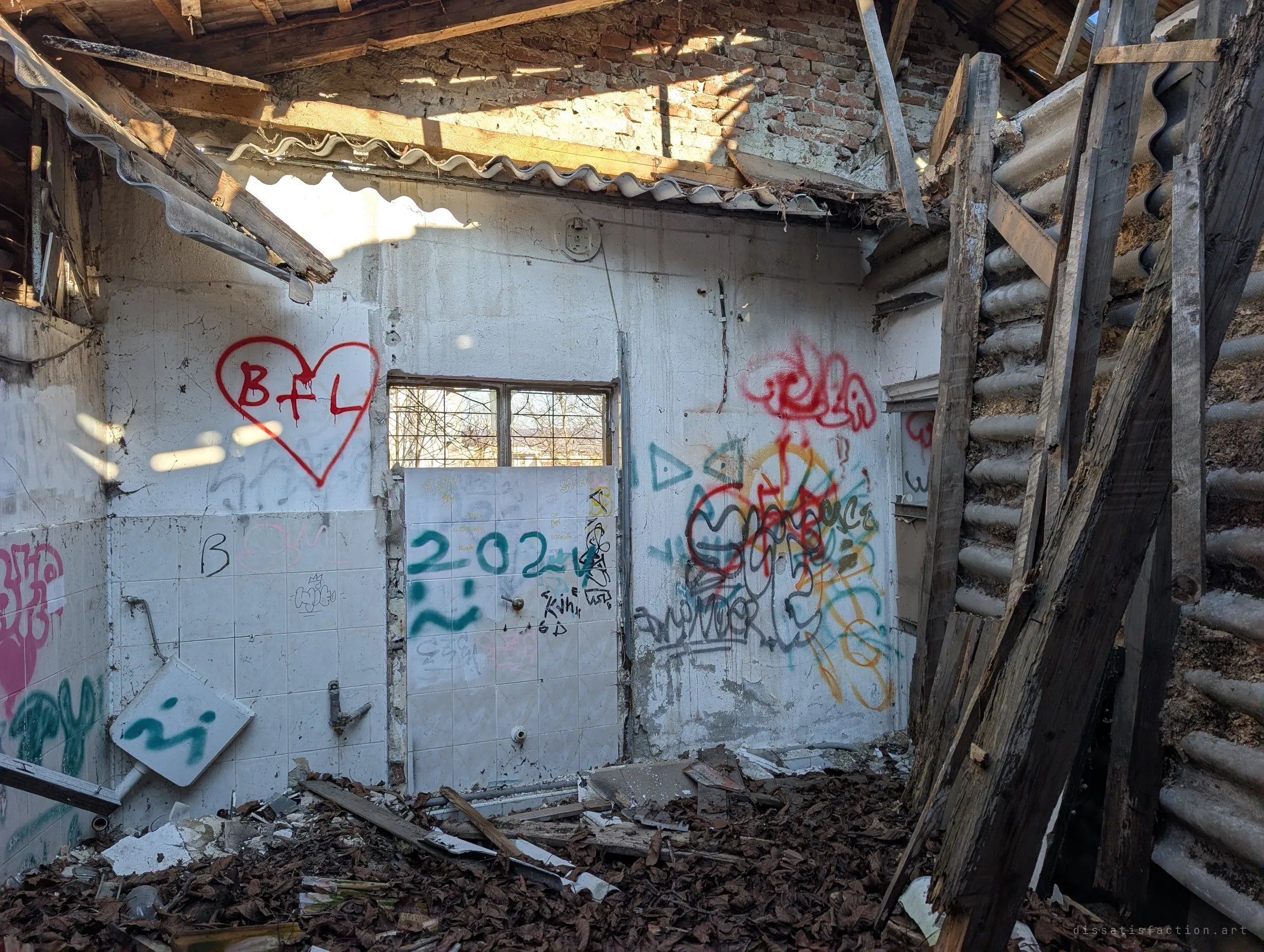 The end wall of the washroom, its white tiles tagged with a red heart and a sprayed year, stands under broken asbestos sheeting and an exposed brick gable. Leaves and planks from the fallen roof cover the floor.