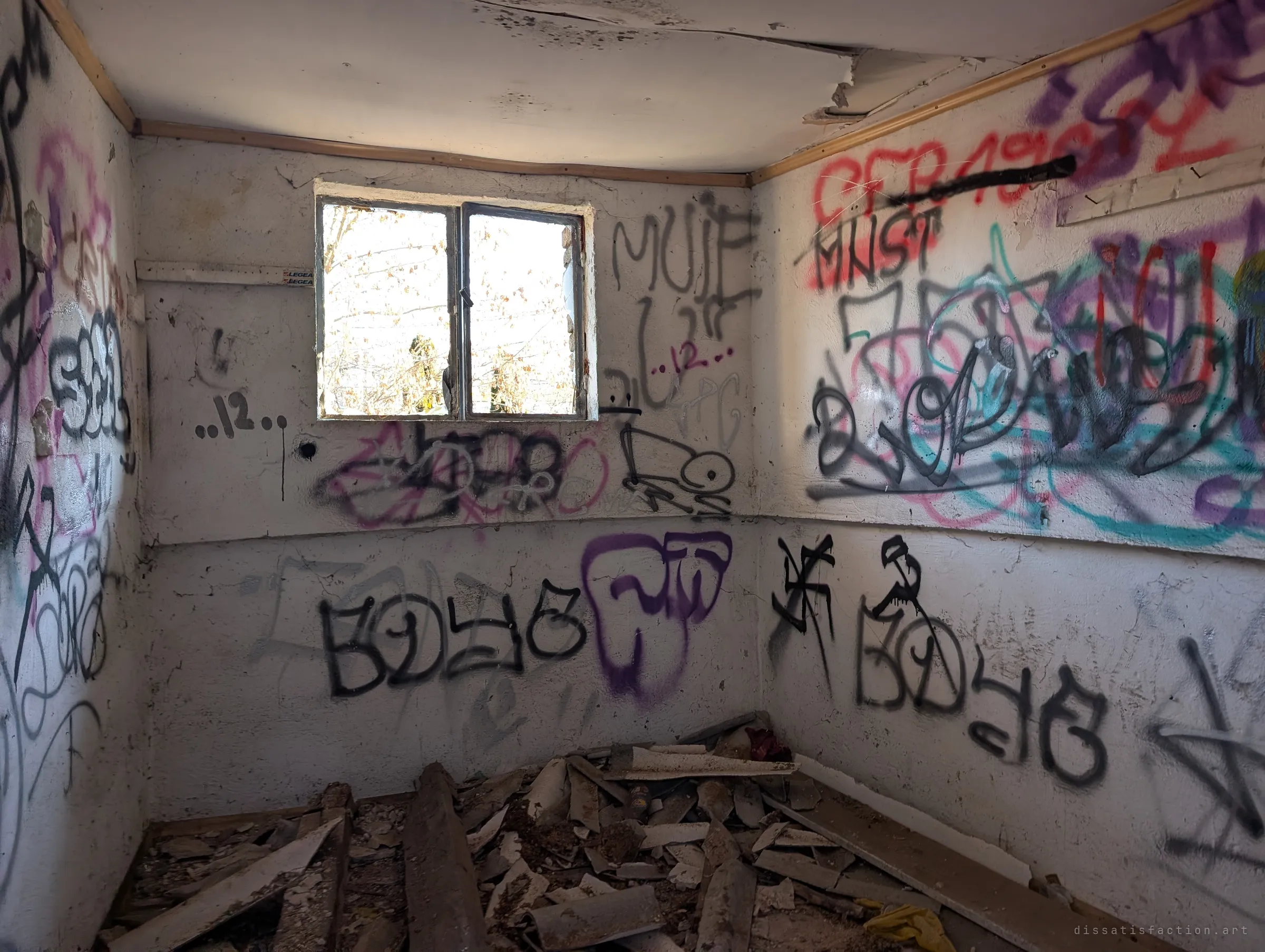 A small whitewashed room with one sunlit window is covered wall to wall in black and coloured tags, with broken roof sheeting heaped in the corner. The low ceiling and bare walls make it feel like a cell.