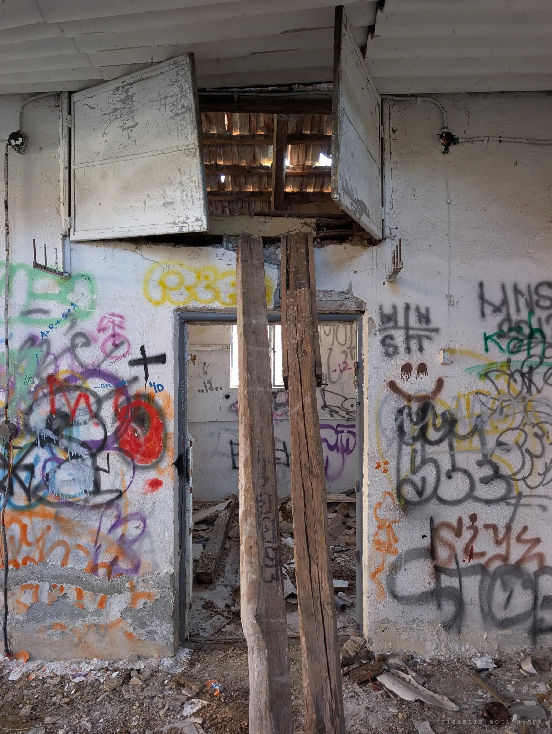 Two rough wooden posts prop up the lintel of a doorway beneath a pair of metal hatches hanging open into the roof space. Through the door is the small tagged room with its single window.