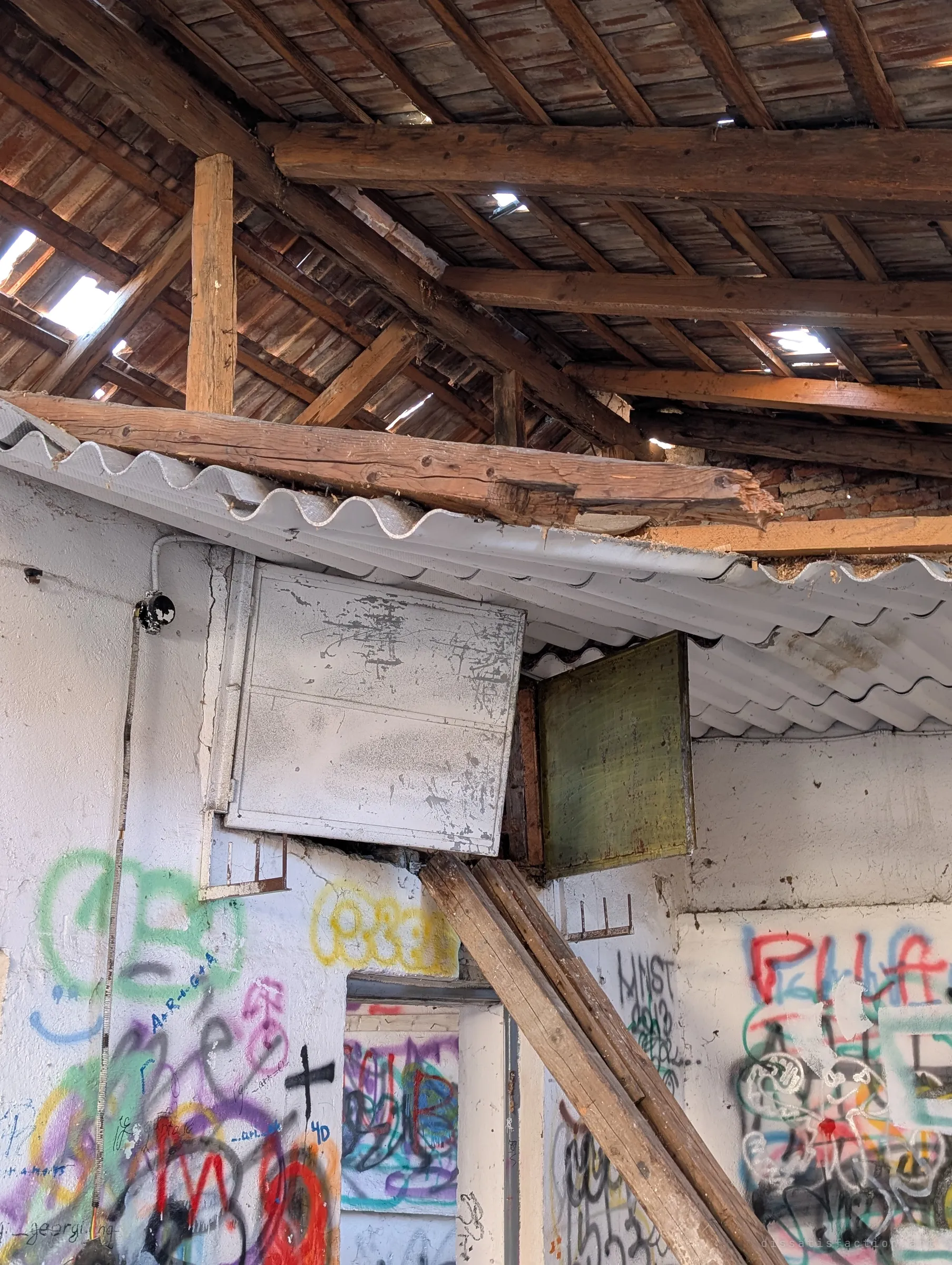 Looking up at the same hatches, the old timber roof shows through, its tiles slipping and letting in shards of light. A snapped beam rests on the corrugated sheeting above the graffiti.