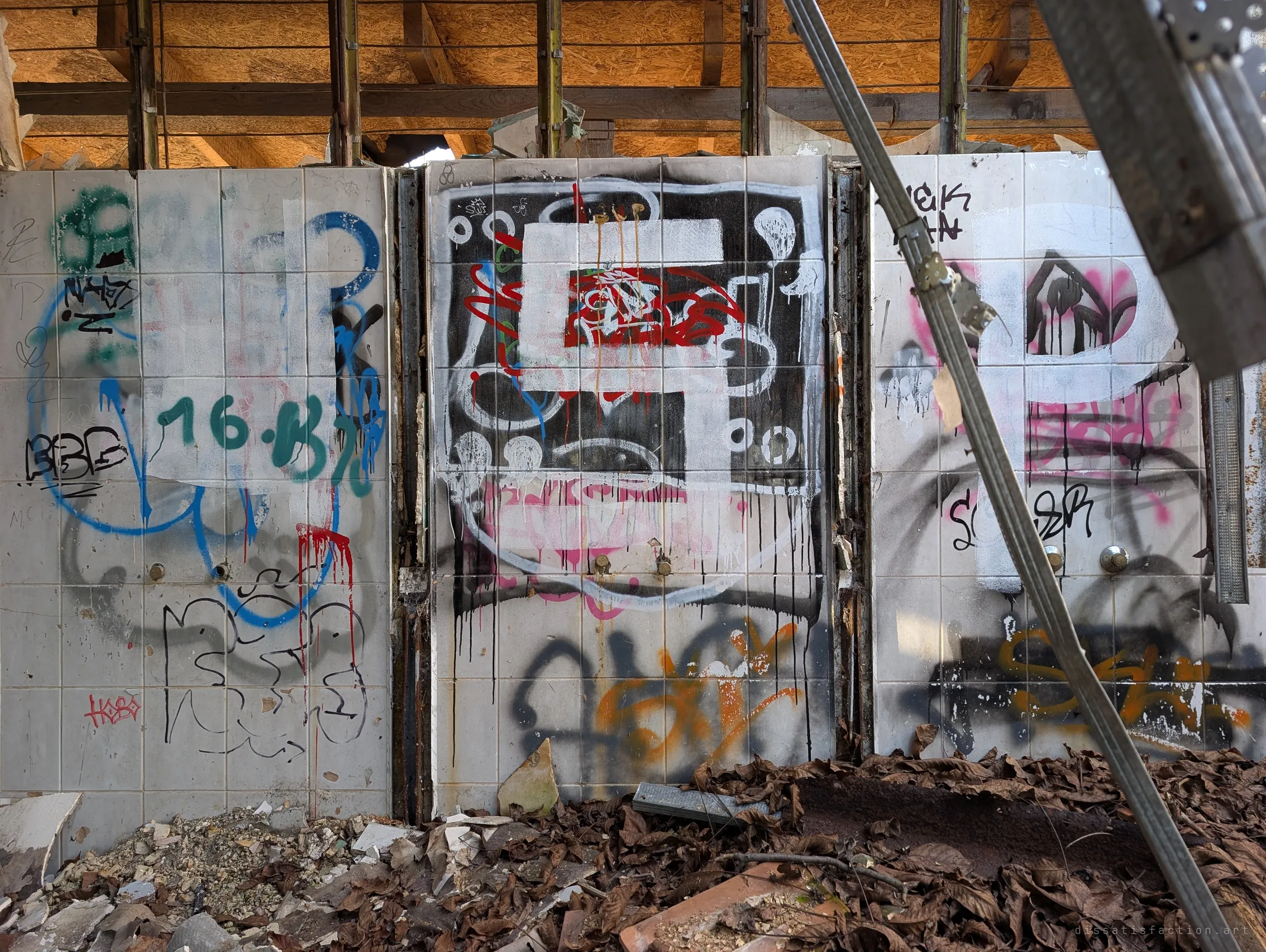 Three tiled shower bays stand side by side, their taps gone and their walls painted over with large graffiti pieces. A fallen metal strut cuts across the frame and dead leaves drift against the base.
