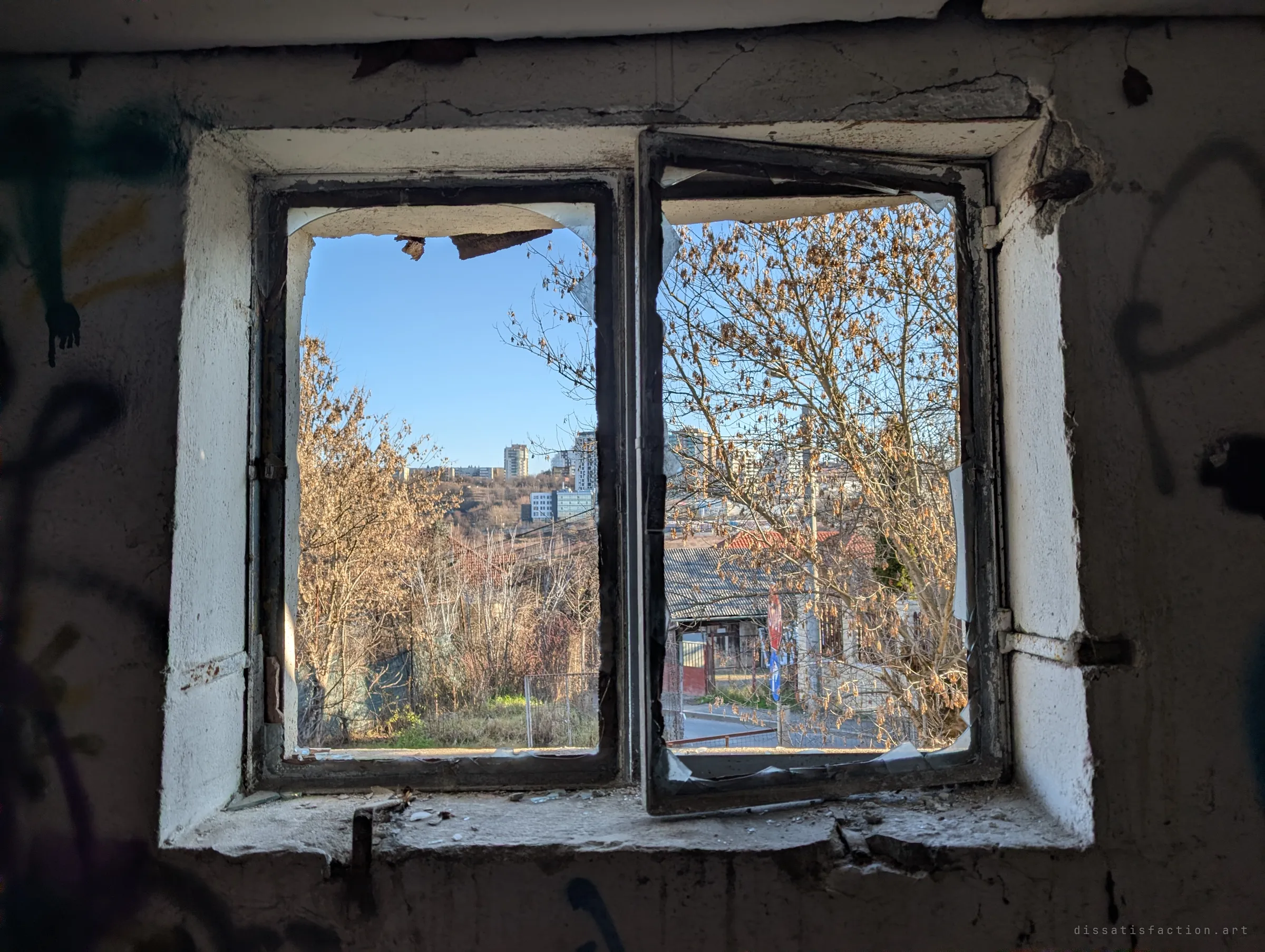A broken double window looks out from the dark interior onto a sunny winter street and tall buildings on a distant hill. The glass hangs in jagged shards from the frame.