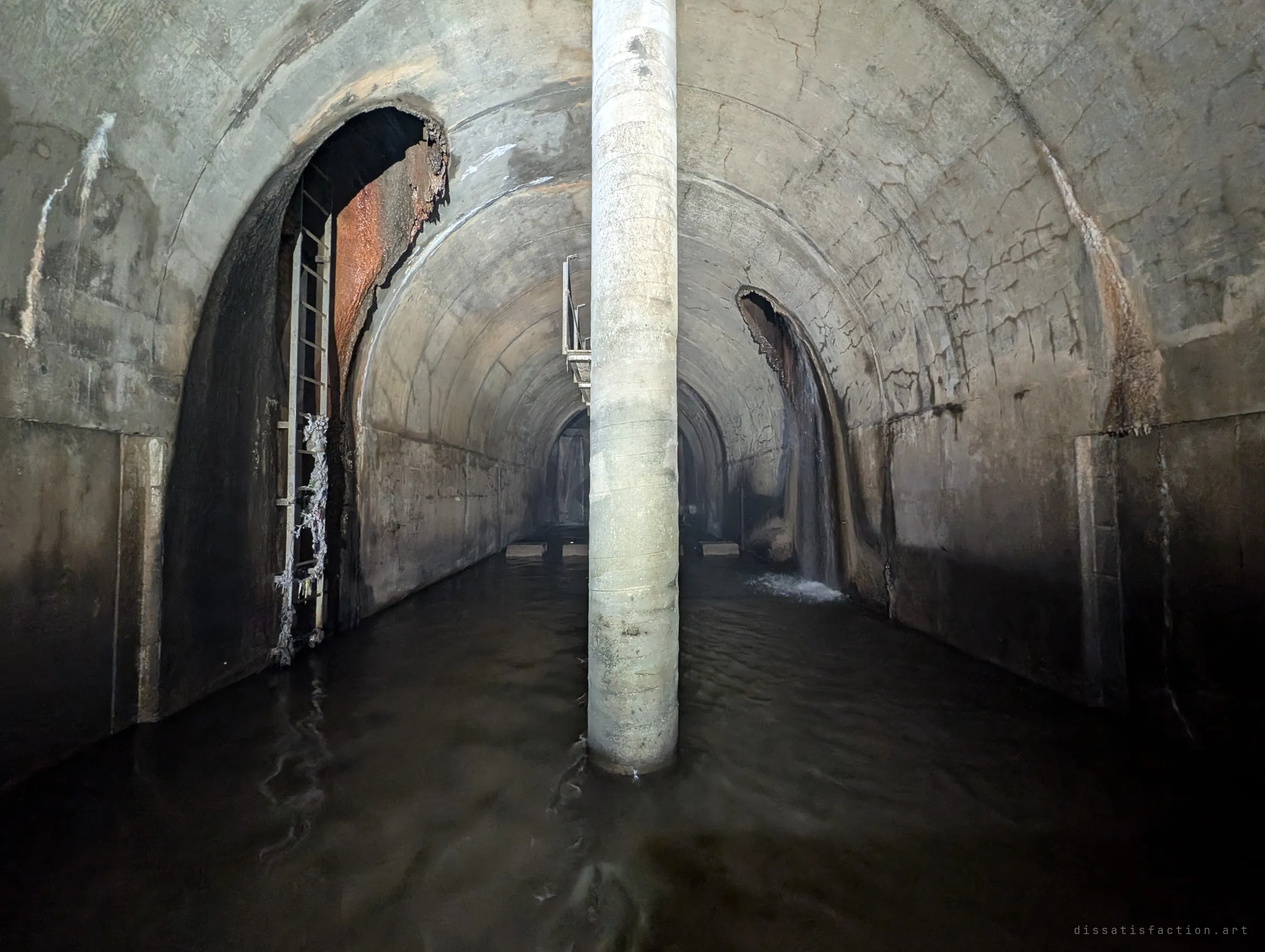 A vaulted underground chamber split by a single round concrete column, dark water on the floor, a ladder climbing into a rust-stained opening on the left and a side inlet pouring a small waterfall on the right.