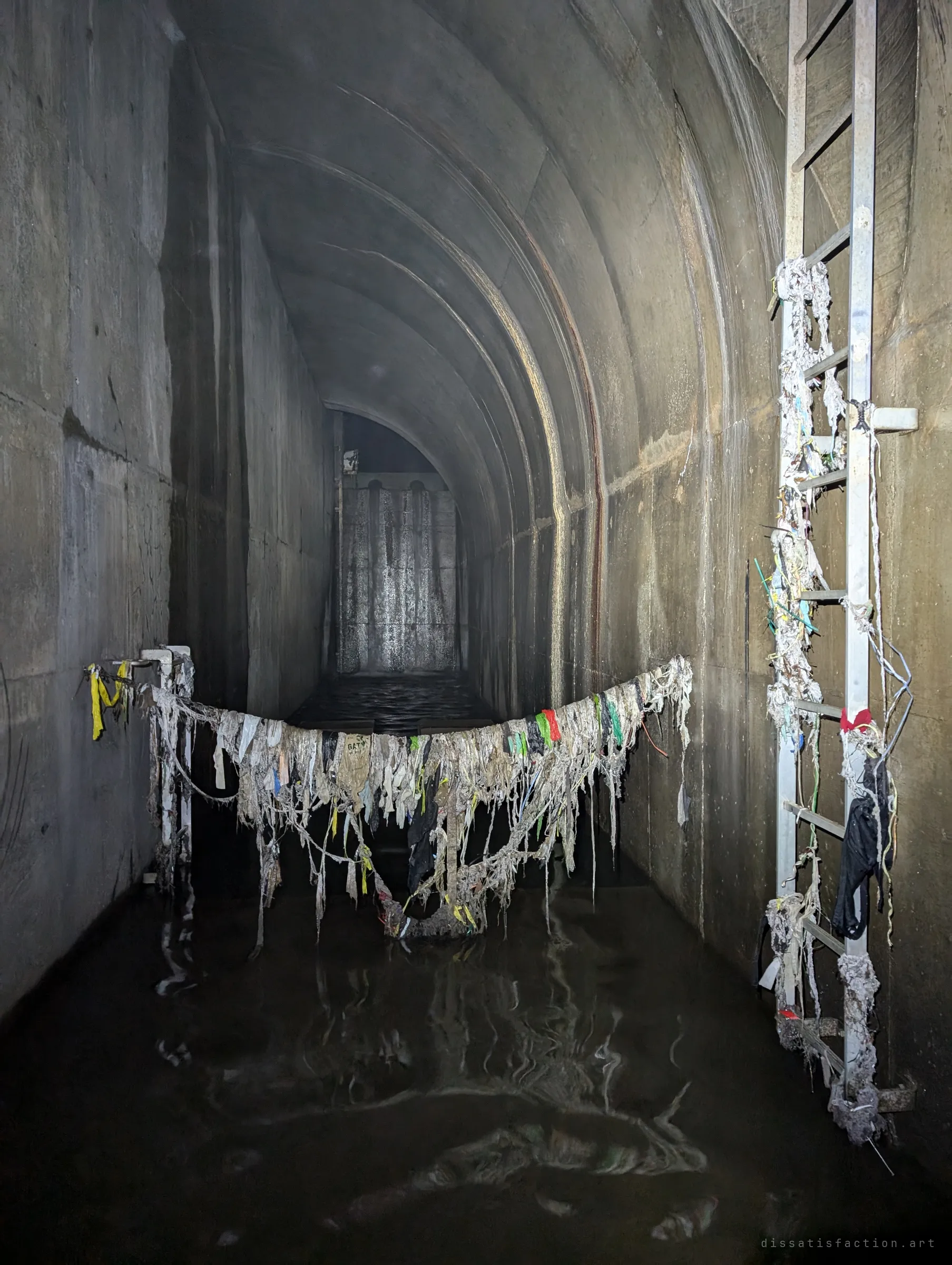 A cable strung across the tunnel sags under a curtain of snagged rags, wipes and plastic, with a debris-wrapped ladder on the right; it shows how high the water rises when it storms.